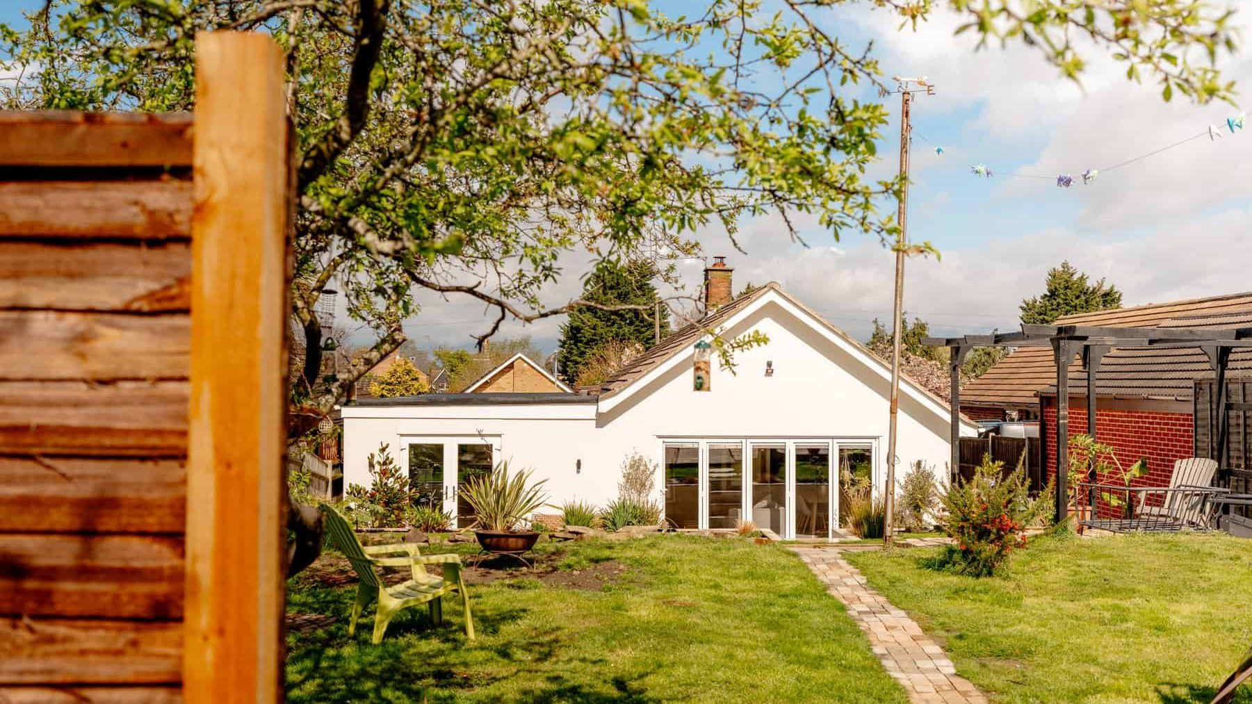 A sunny backyard with a pathway leading to a white single-story house with french doors. trees and a wooden fence surround the well-maintained garden area.