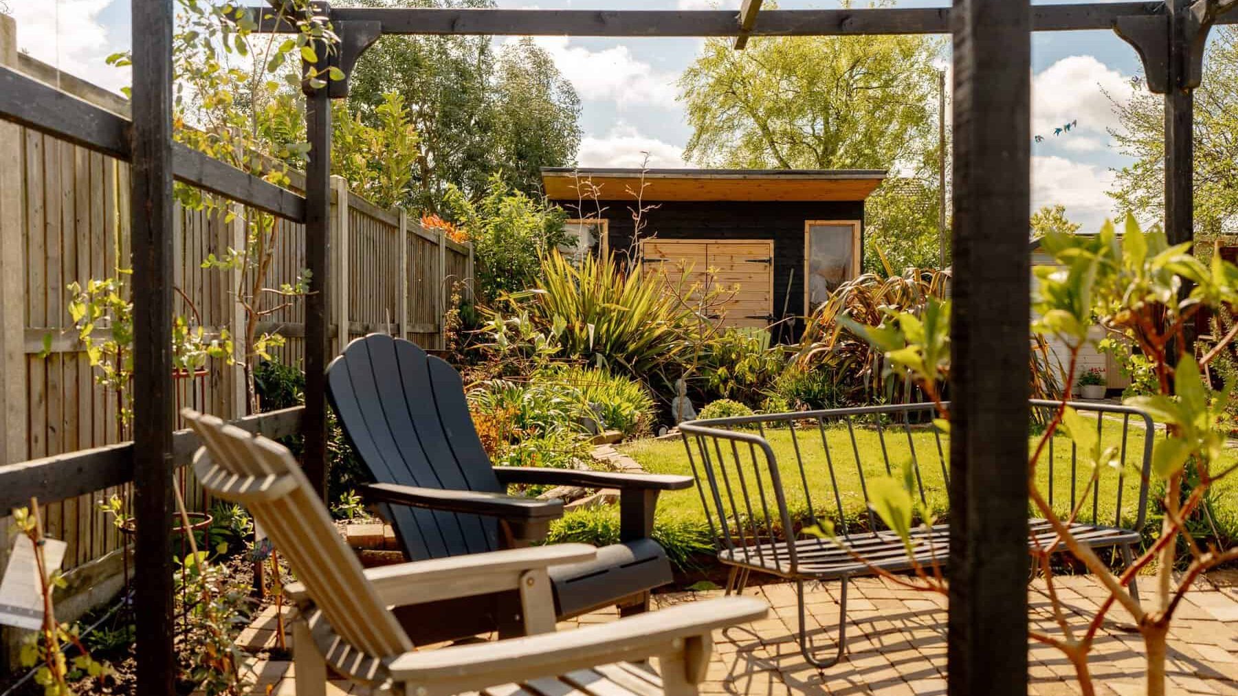 A cozy garden with a wooden pergola, an adirondack chair, and lush greenery. a well-kept lawn leads to a small wooden shed surrounded by plants under a sunny sky.