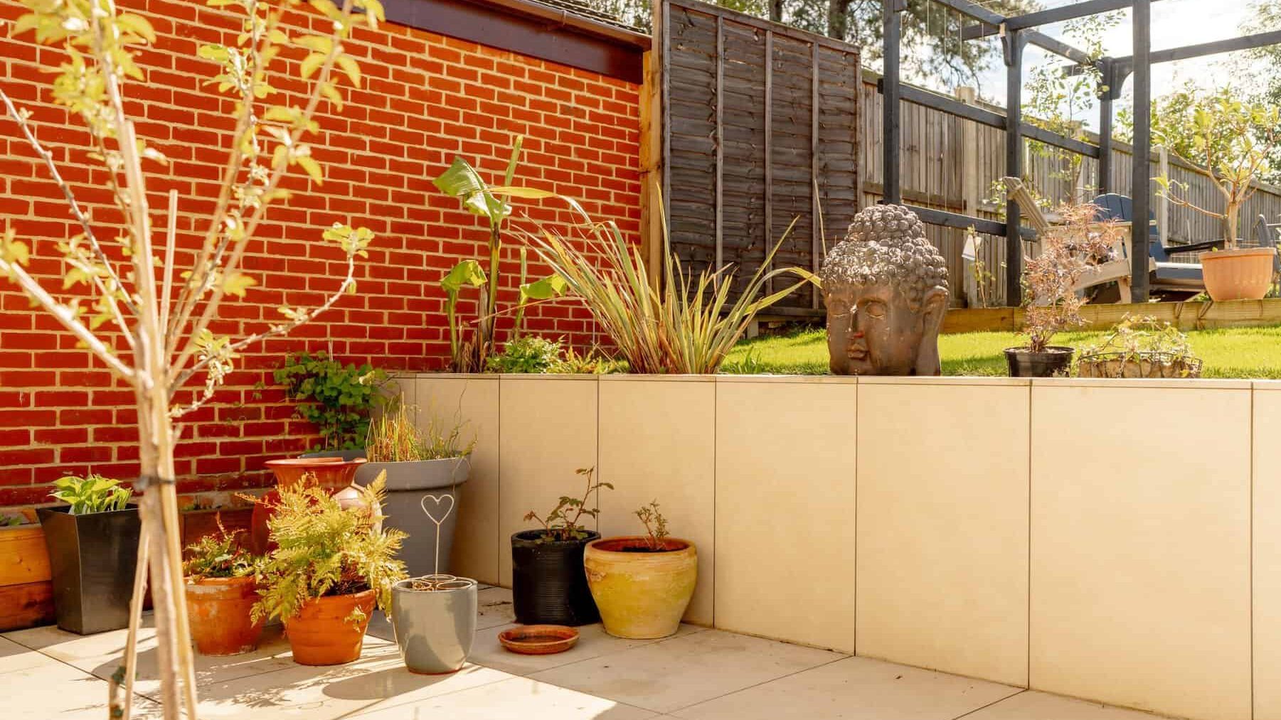 A tranquil garden corner featuring a buddha head statue, surrounded by potted plants and a tall brick wall. sunlight brightens the area, highlighting various textures and colors in the serene setting.