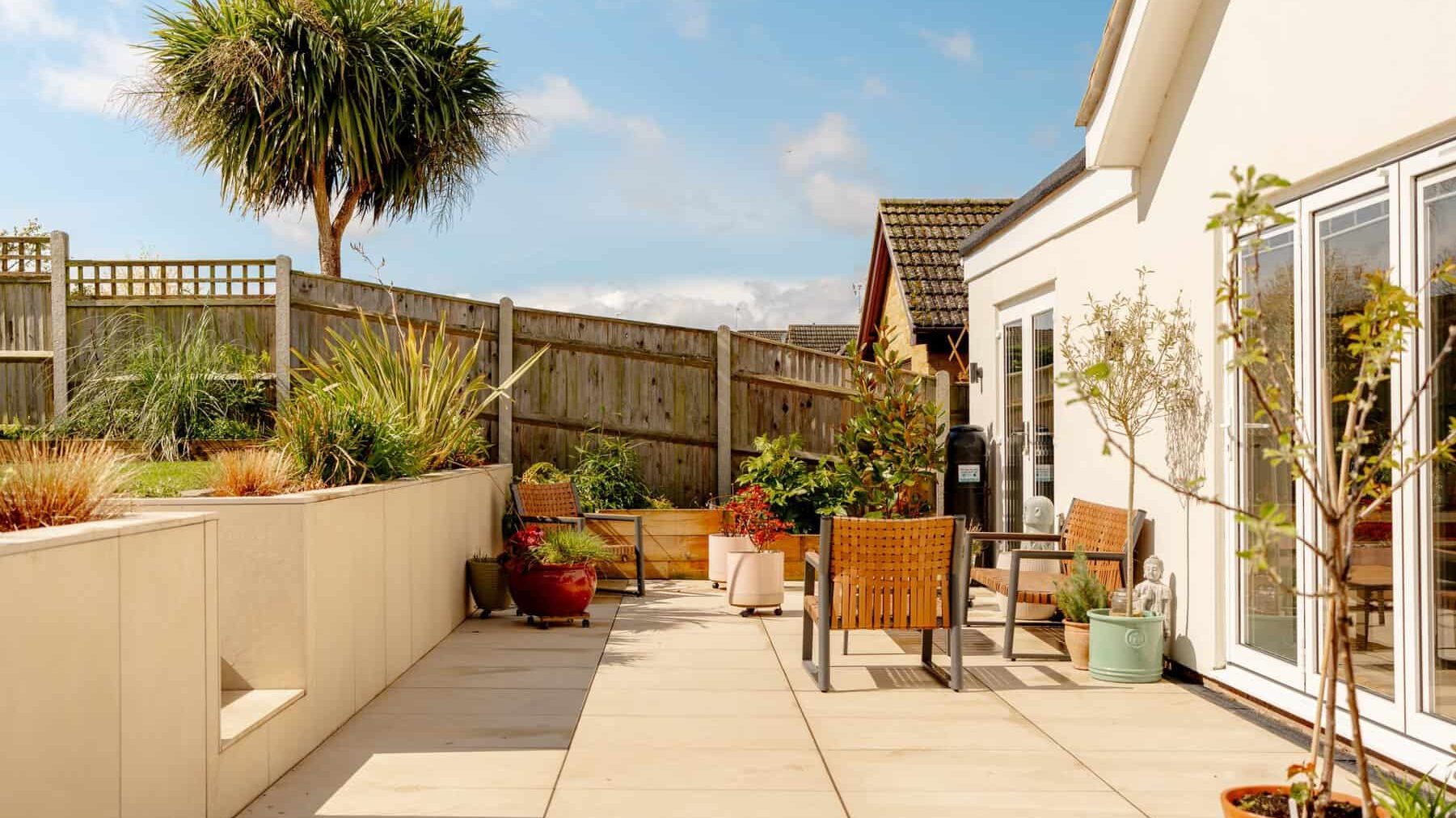 A sunny rooftop garden featuring potted plants, a small tree, and wooden furniture. wooden fencing surrounds the area and a clear blue sky is visible above.