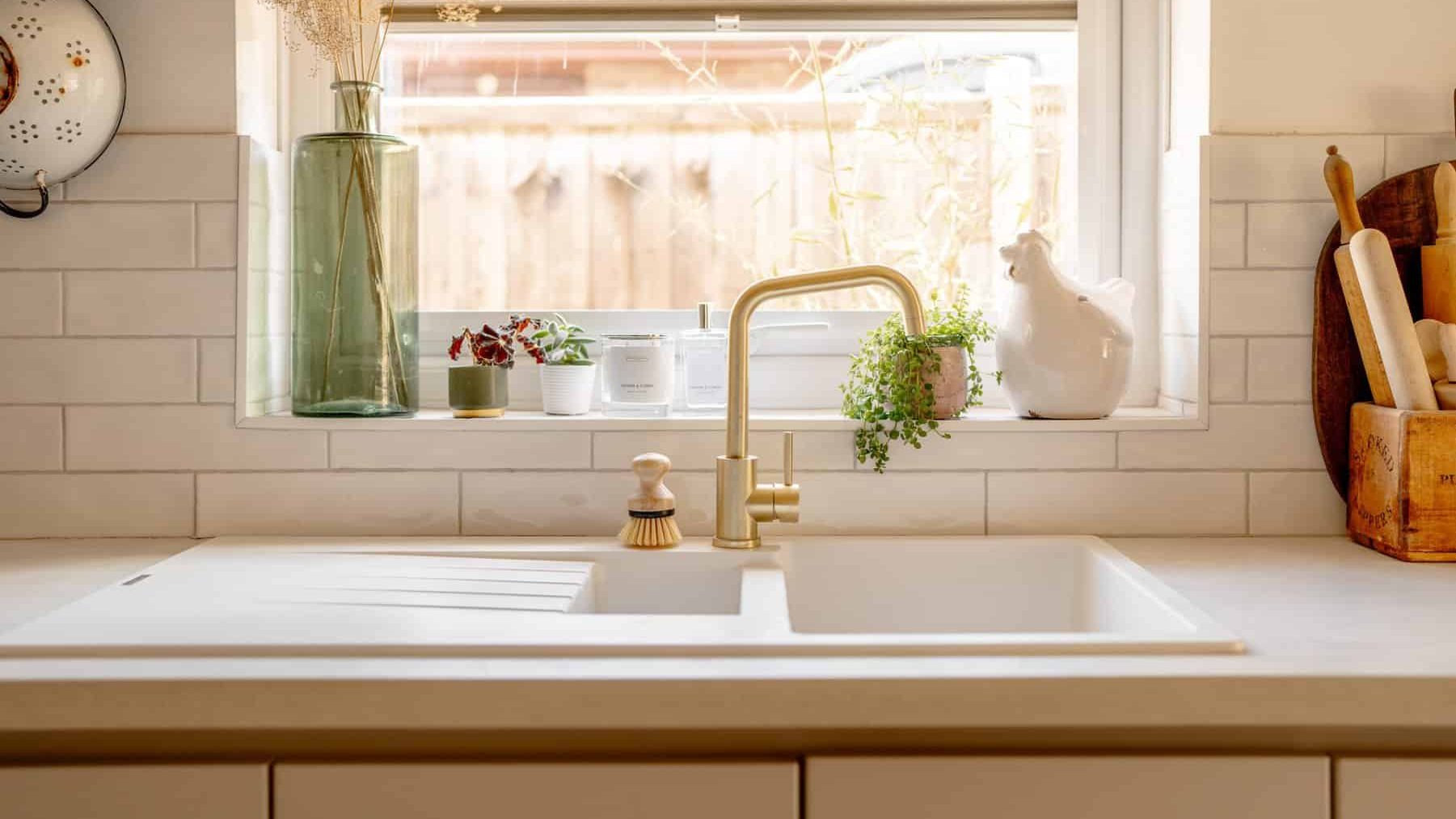 A bright kitchen sink area with white countertops and gold faucet, featuring decorative items like a rooster figurine and potted plants by a window with sunlight streaming in.