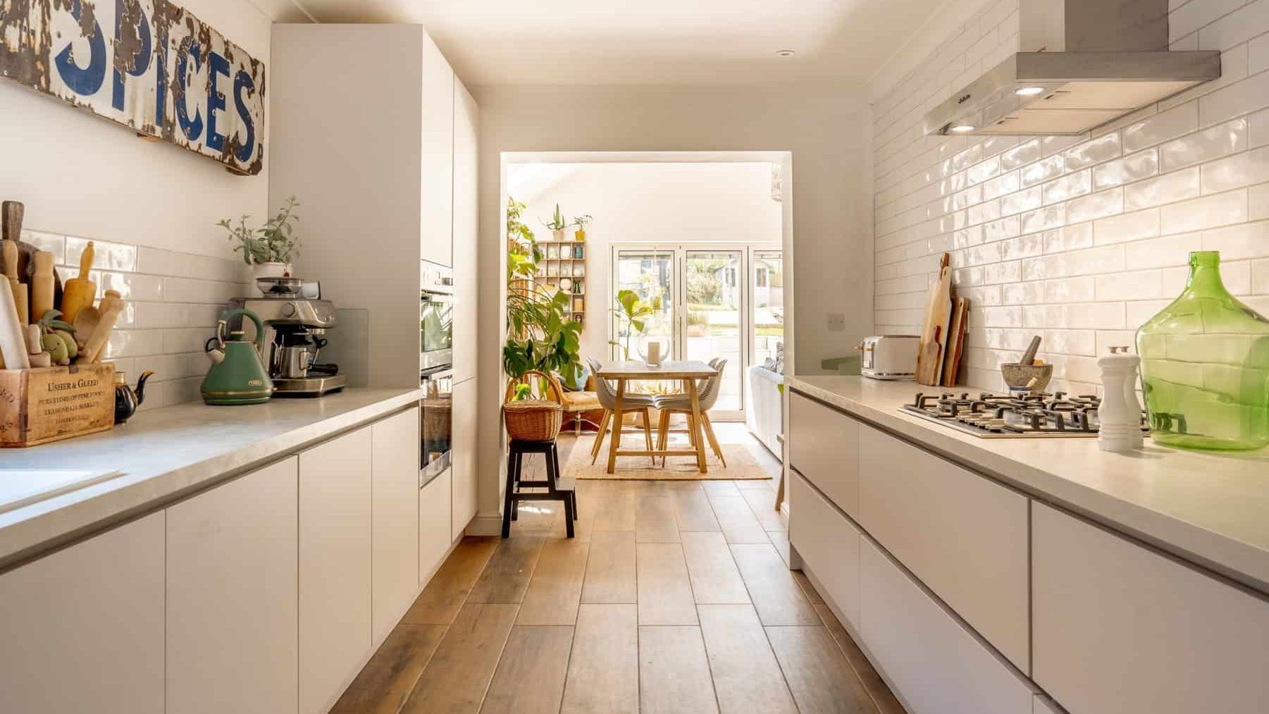 A bright, modern kitchen featuring white cabinetry, wooden flooring, and a central aisle. the room opens into a sunny dining area with a round table and chairs, and decorative items are placed throughout.