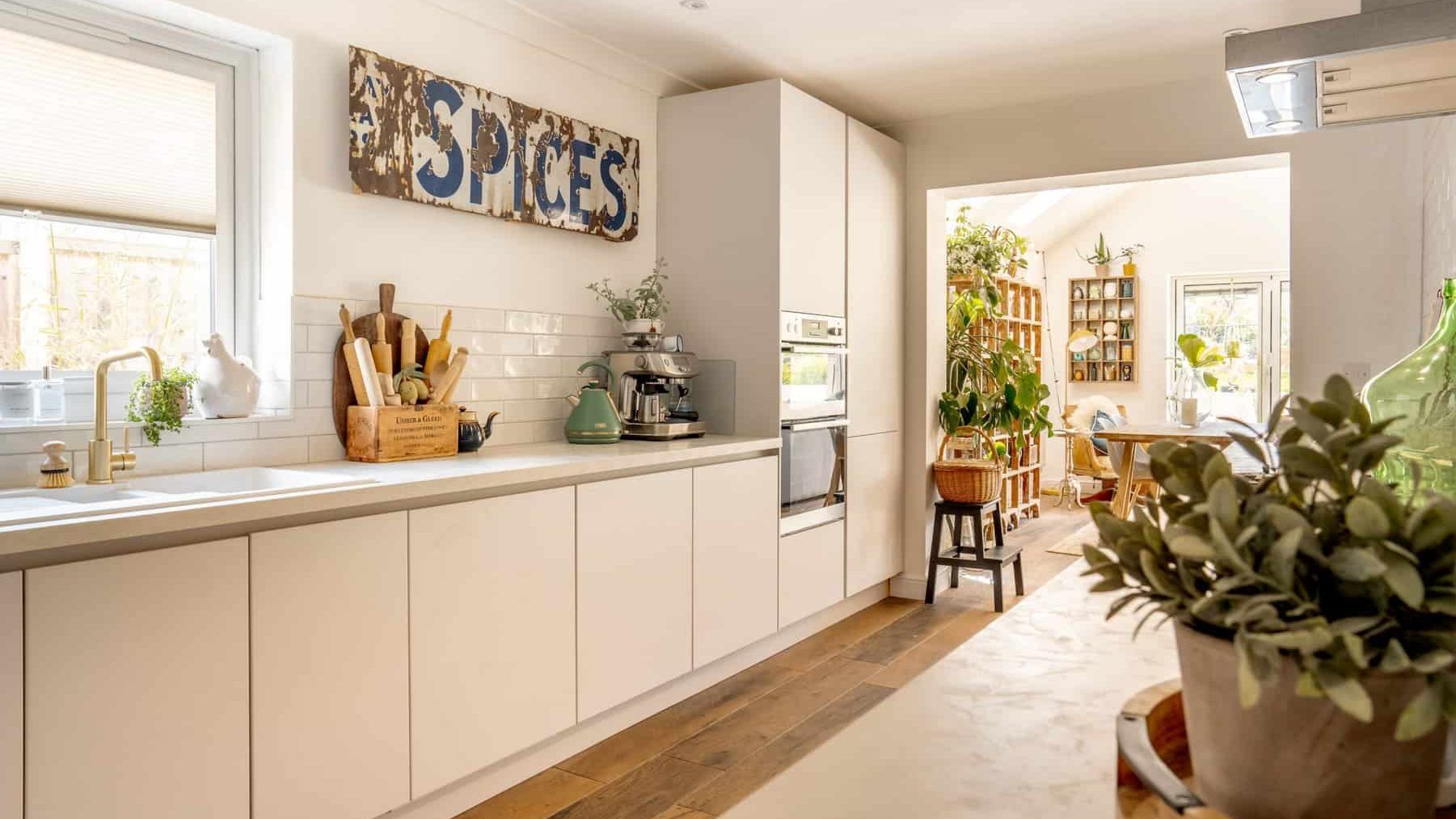 A modern kitchen with white cabinetry and a long counter. features include a “spices” sign on the wall, a wooden table with plants, and sunlit windows.