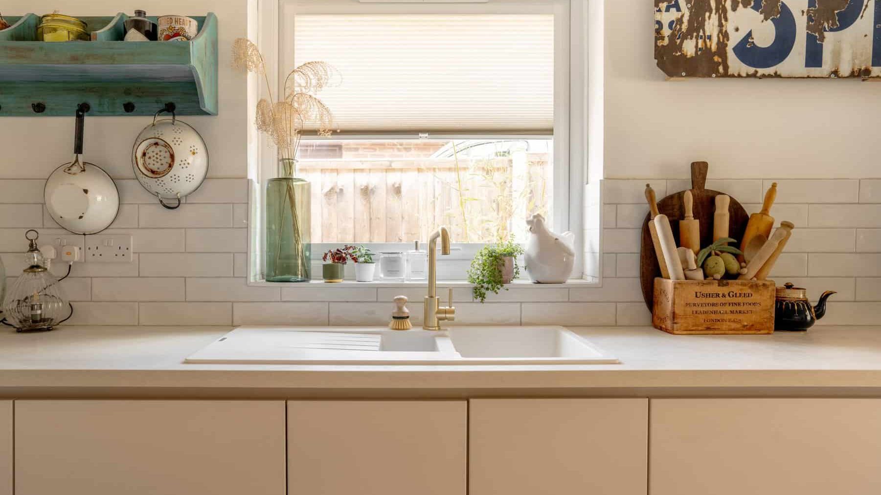 A bright kitchen interior with a clean white countertop featuring a sink beneath a window, surrounded by various kitchen utensils and decorative items. the overall decor includes a neutral palette with wood accents.