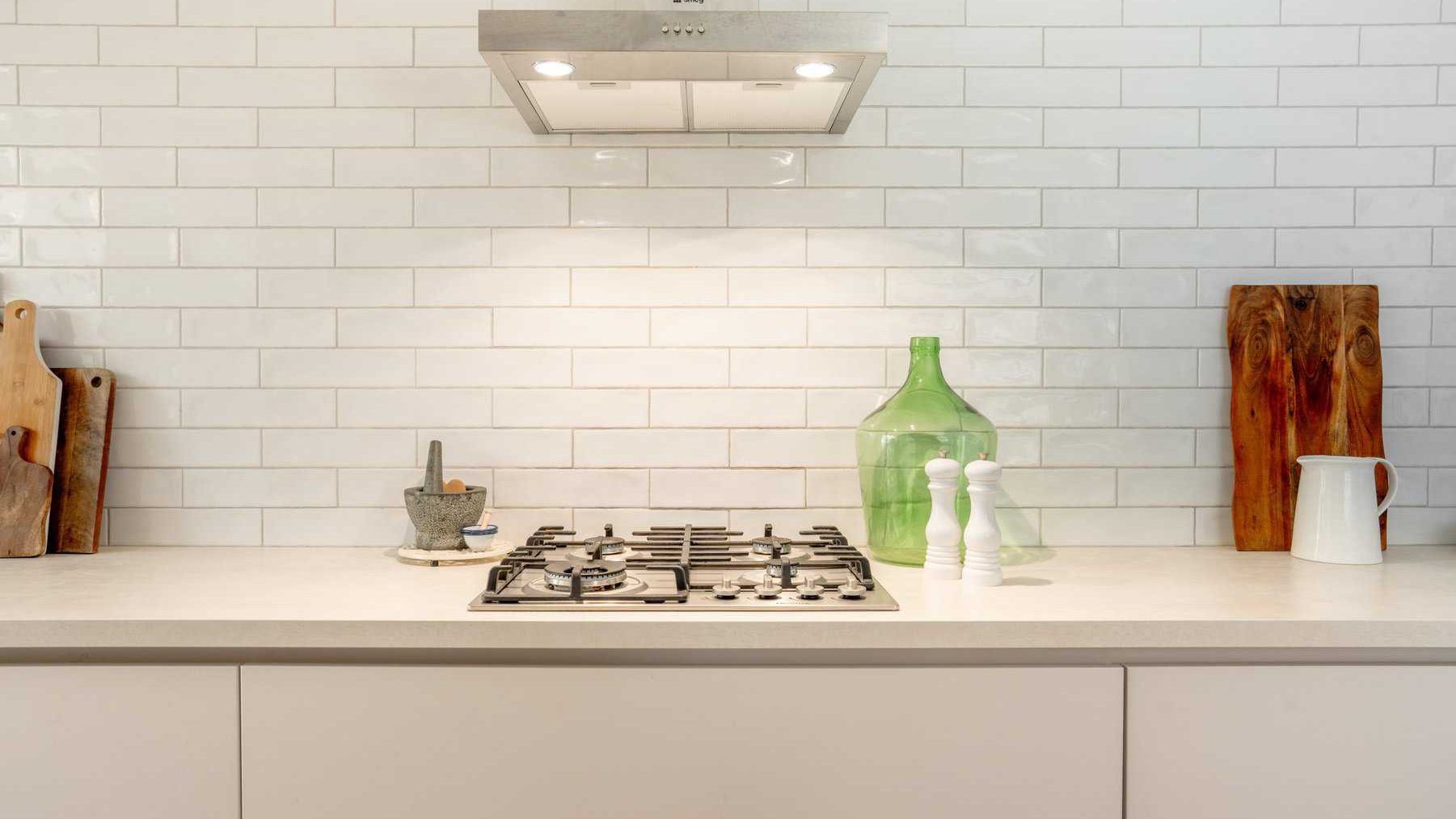 A modern kitchen counter featuring a gas stove, a range hood, and various decorative items including a large green bottle, wooden cutting boards, and a white jug against a white subway tile backsplash.