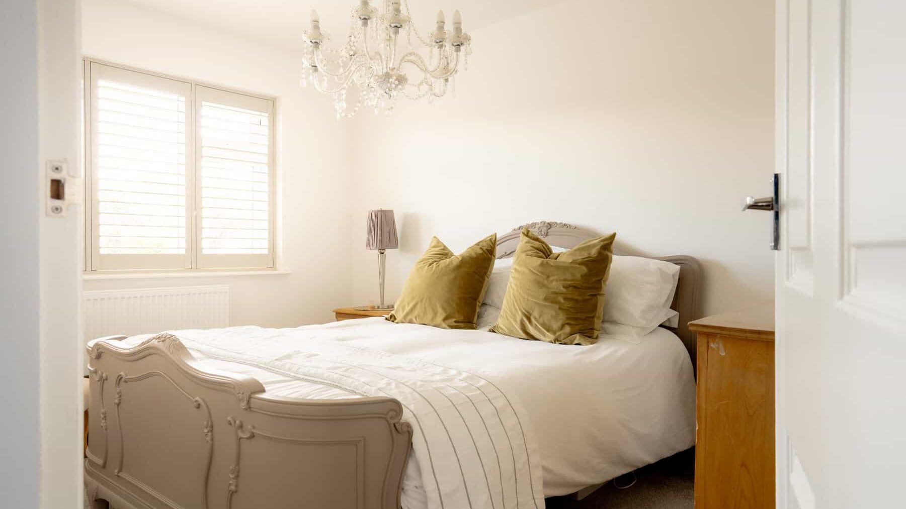 A neatly organized bedroom featuring a large double bed with a white duvet and olive green pillows. the room includes a classic wooden dresser, a bedside lamp, and a chandelier, with natural light coming through a window with blinds.
