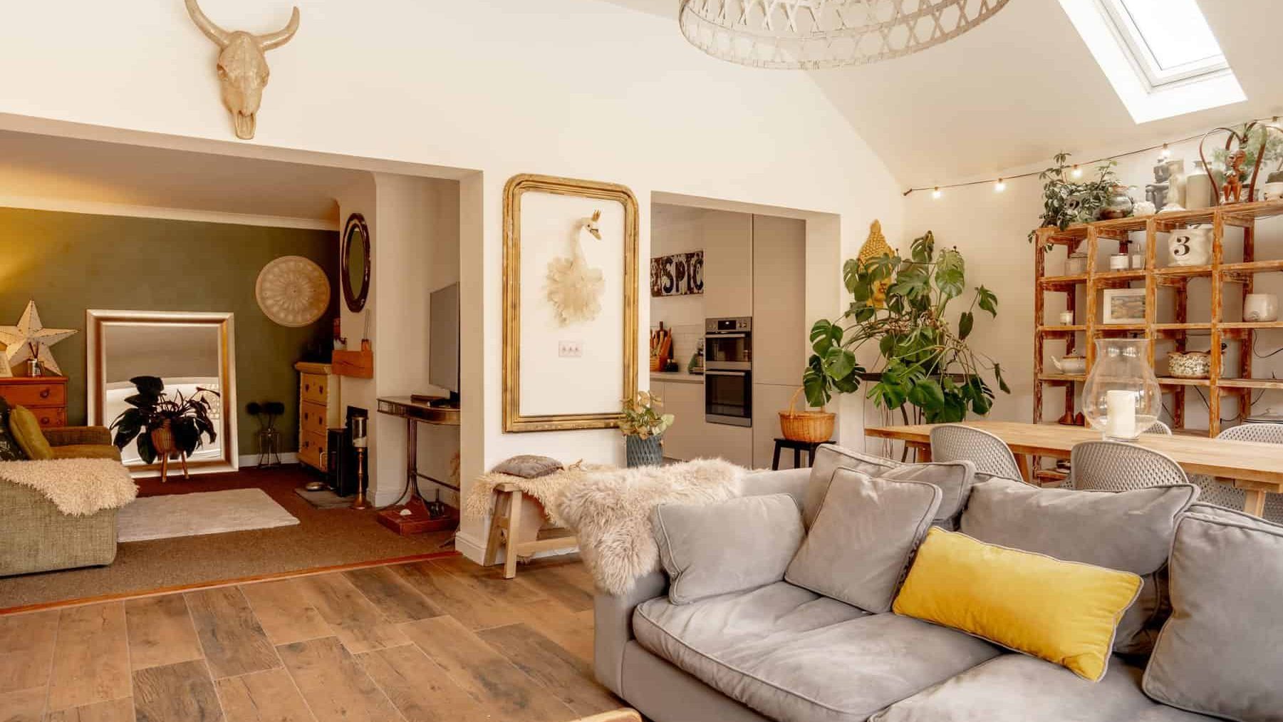 A cozy living room featuring a gray sectional sofa with yellow pillows, wooden flooring, a skylight, and decorative items including a large mirror, plants, and wall-mounted art.