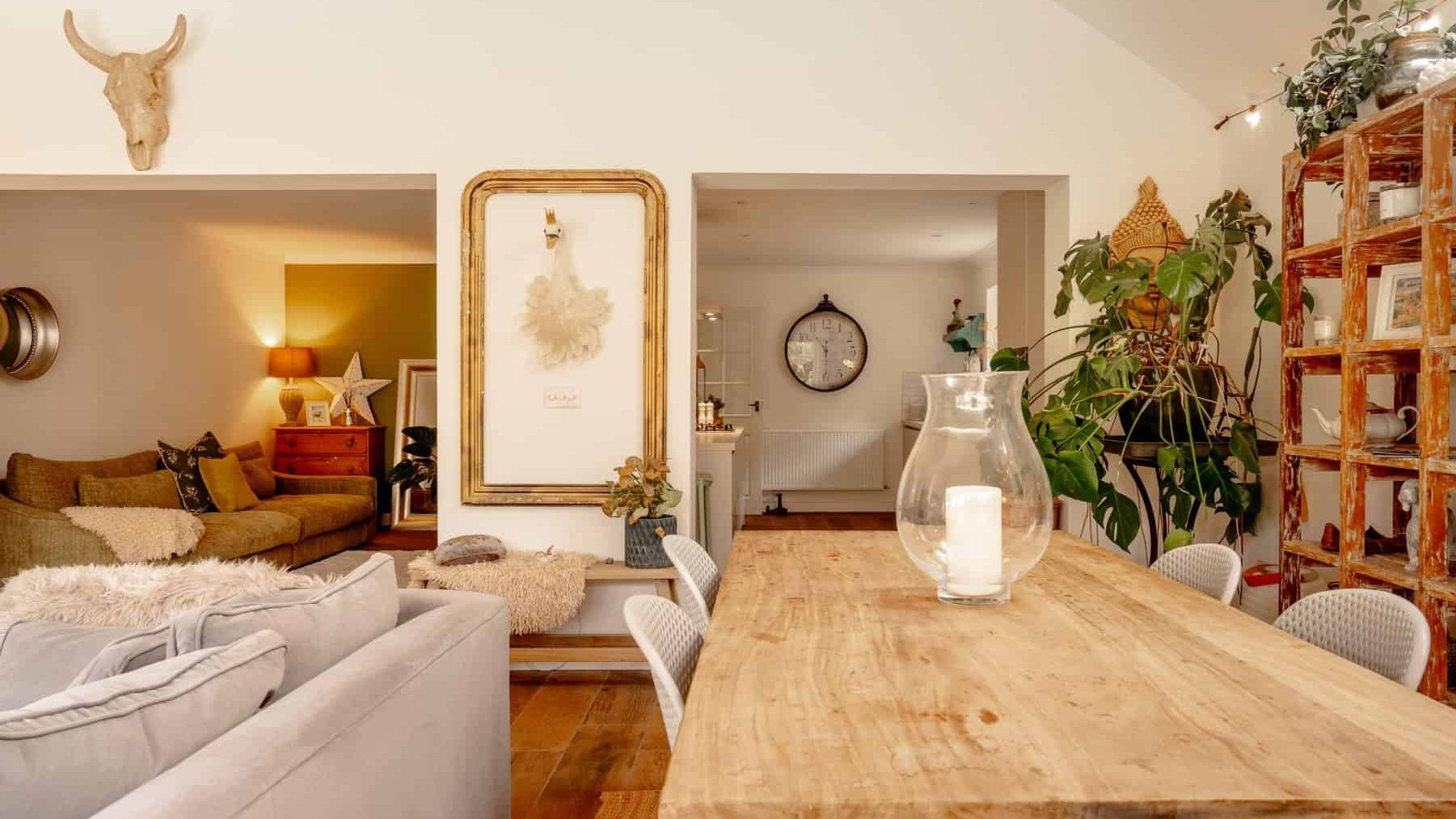 A cozy, warmly lit living room with a rustic wooden dining table in the foreground, plush sofas, decorative plants, and a stylish, inviting open layout leading to adjacent rooms.