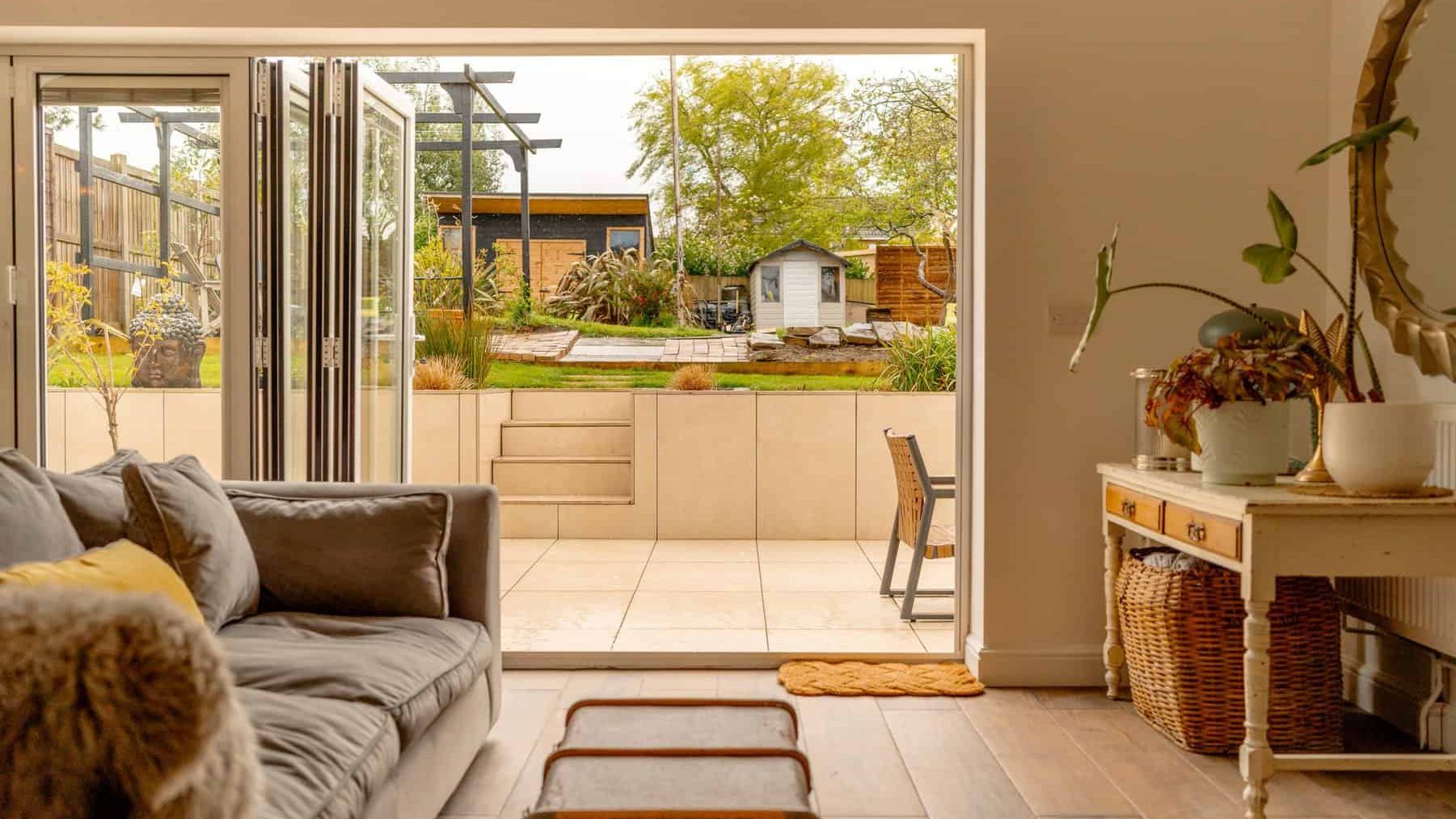 A cozy living room opens to a garden through sliding glass doors, featuring a plush sofa, indoor plants, and a view of a well-maintained yard with a shed.