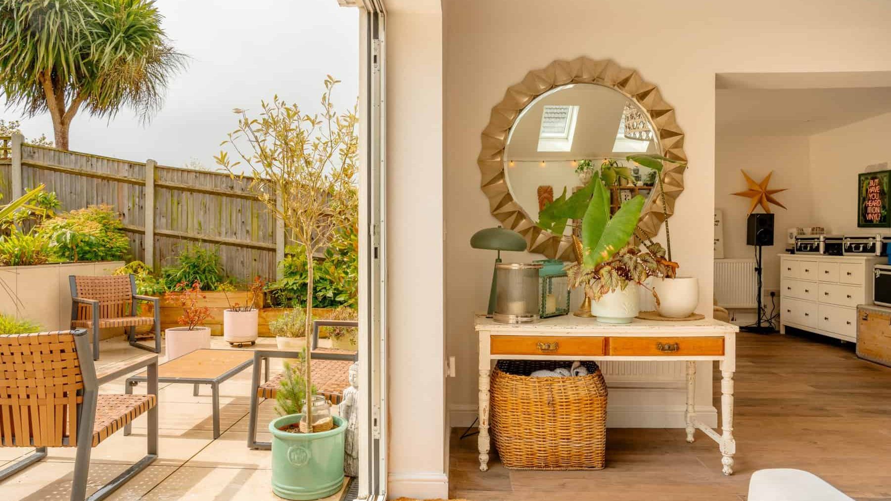 Open patio door leading to a cozy garden with wooden furniture and potted plants. inside, a stylish room features a reflective mirror, decorative items, and a kitchen area in the background.