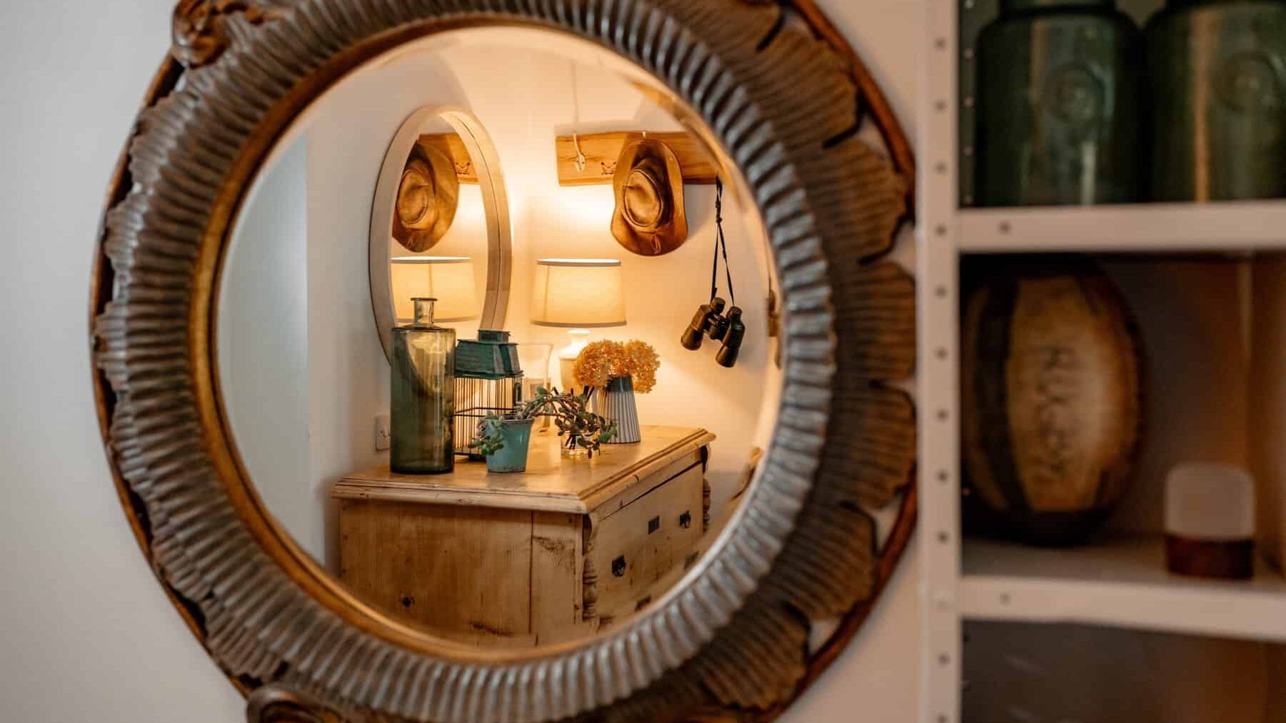 A cozy, well-lit corner reflected in a round, ornate mirror, featuring a small wooden cabinet topped with a vase, books, and a lamp, alongside wall-mounted lights and decorative items on shelves.