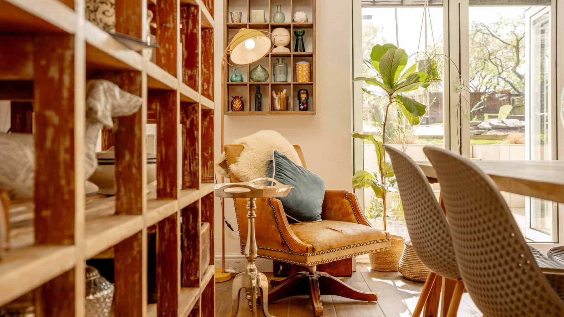 A cozy reading nook by a large window with a leather armchair and throw pillows. a small round table, bookshelves filled with various items, and two chairs in the foreground add to the room's warm ambiance.
