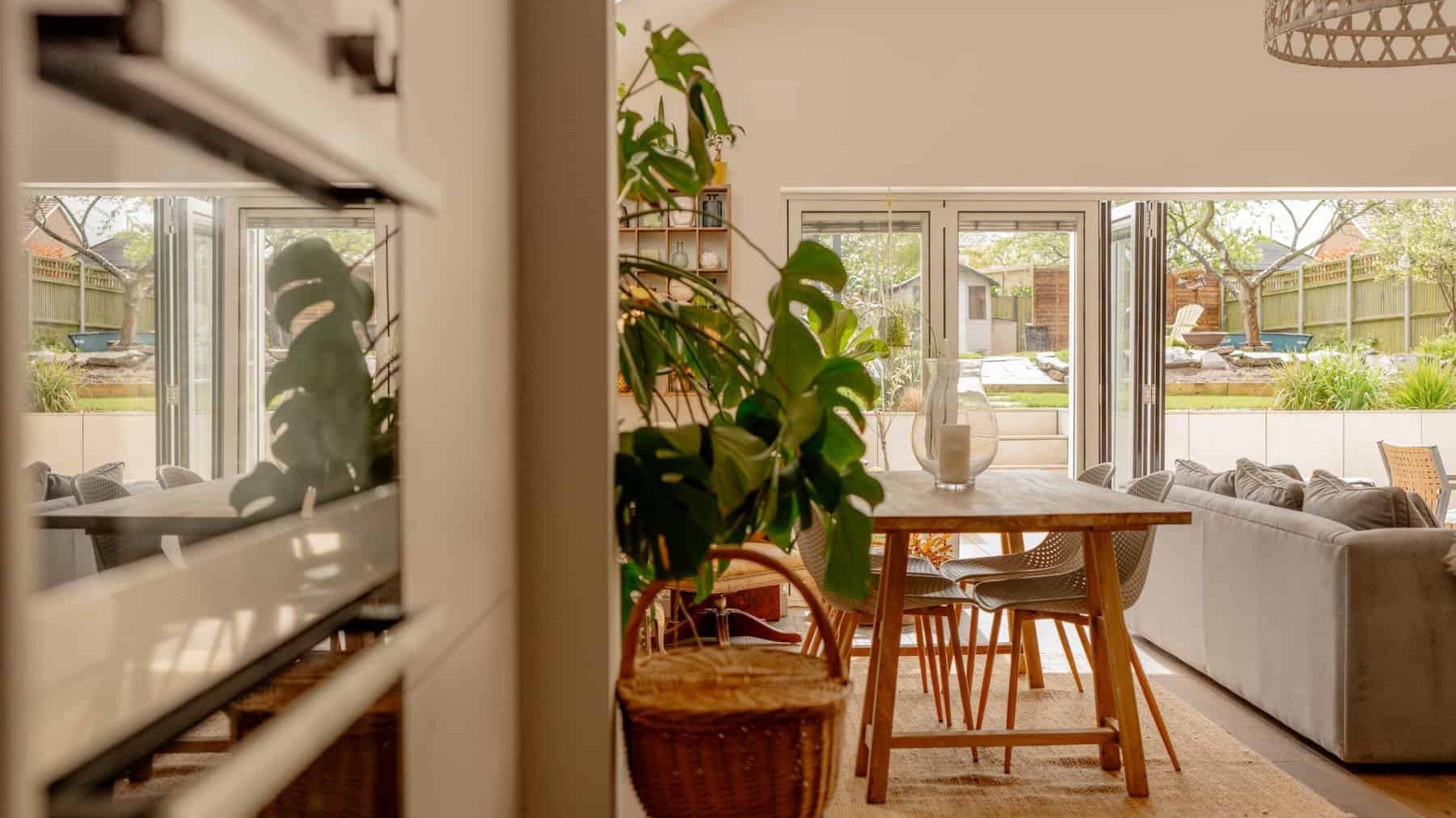 View from a hallway into a sunny dining room with a wooden table, chairs, and a couch. large windows show a garden outside. plants and a wicker basket are also visible.