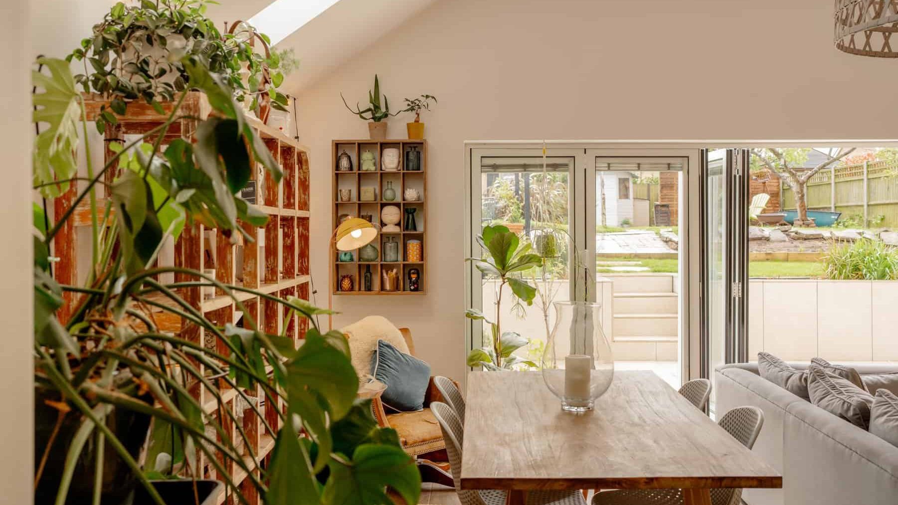 A bright, modern living room with abundant plants, a wooden dining table, comfortable seating, skylights, and open french doors leading to a garden.