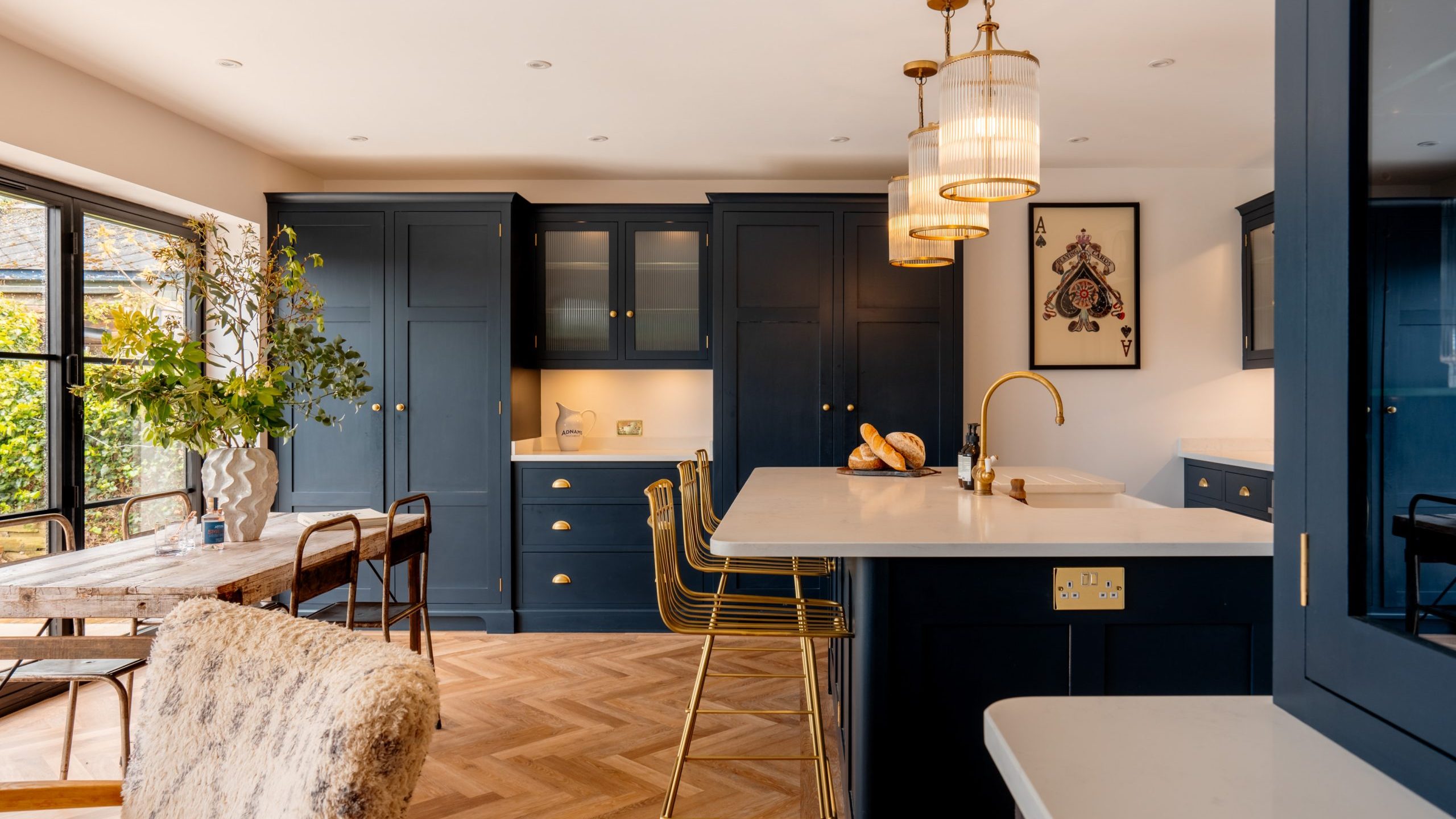 Modern kitchen with dark blue cabinets, a white marble countertop, and a central island. there are gold accents, hanging lights, and a herringbone wood floor. a dining area is visible in the background.
