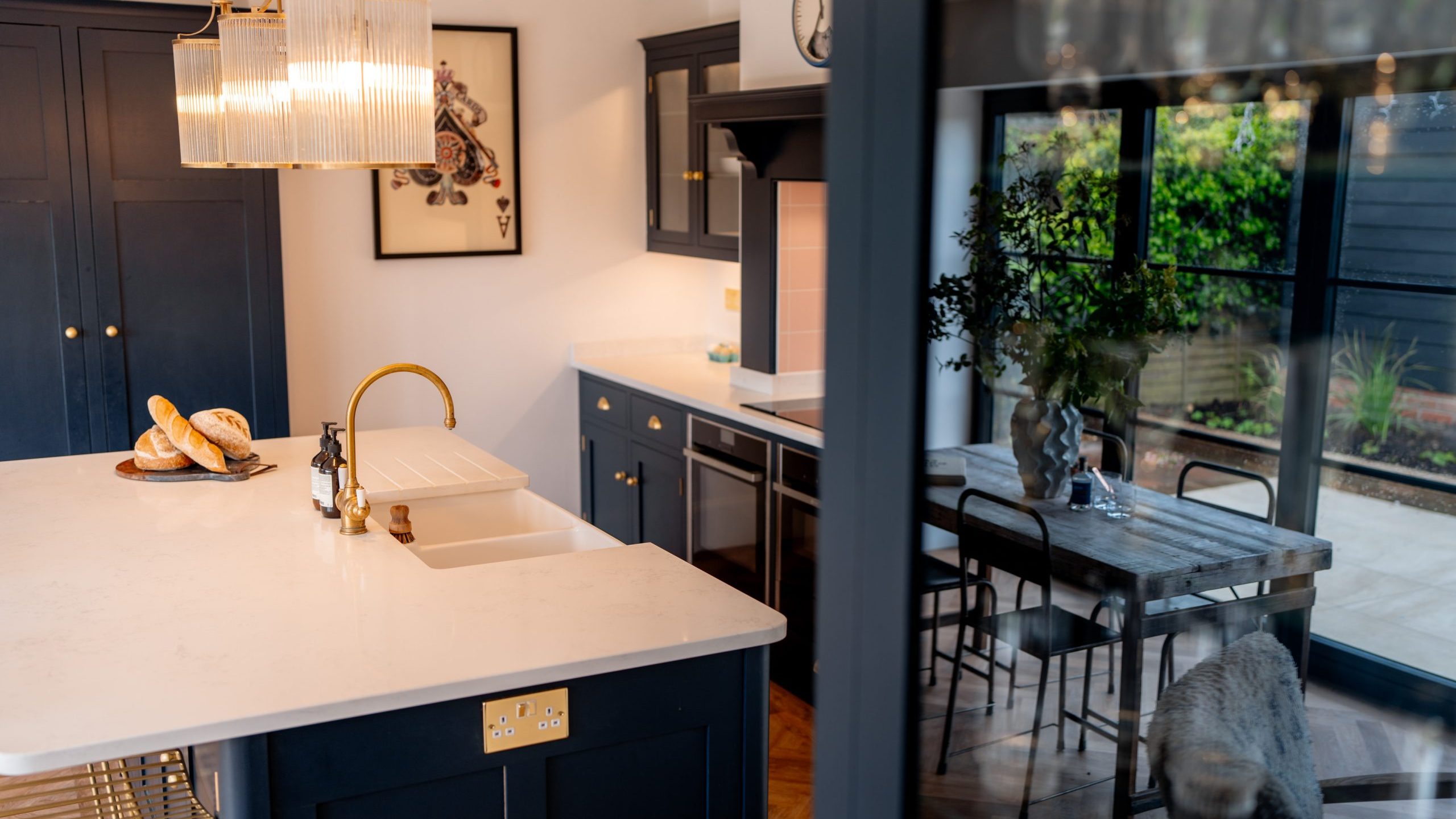 Modern kitchen with blue cabinetry and white countertops, featuring gold fixtures and a marble island. a chandelier hangs above. view through doorway shows a dining area and garden.
