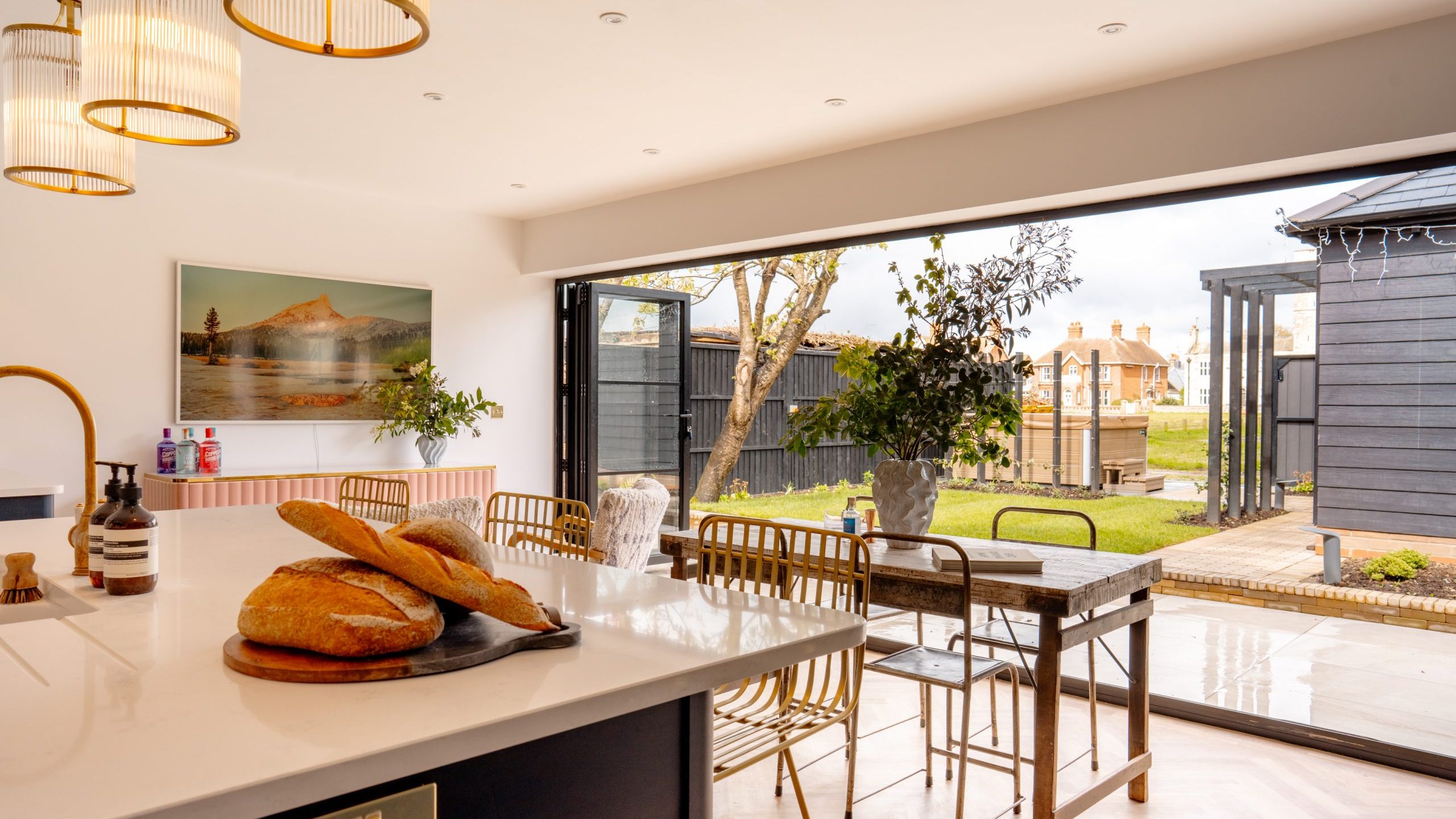 A modern kitchen with a large island featuring freshly baked bread, leading to a dining area with wooden furniture. large glass doors open to a tidy backyard with a garden shed.