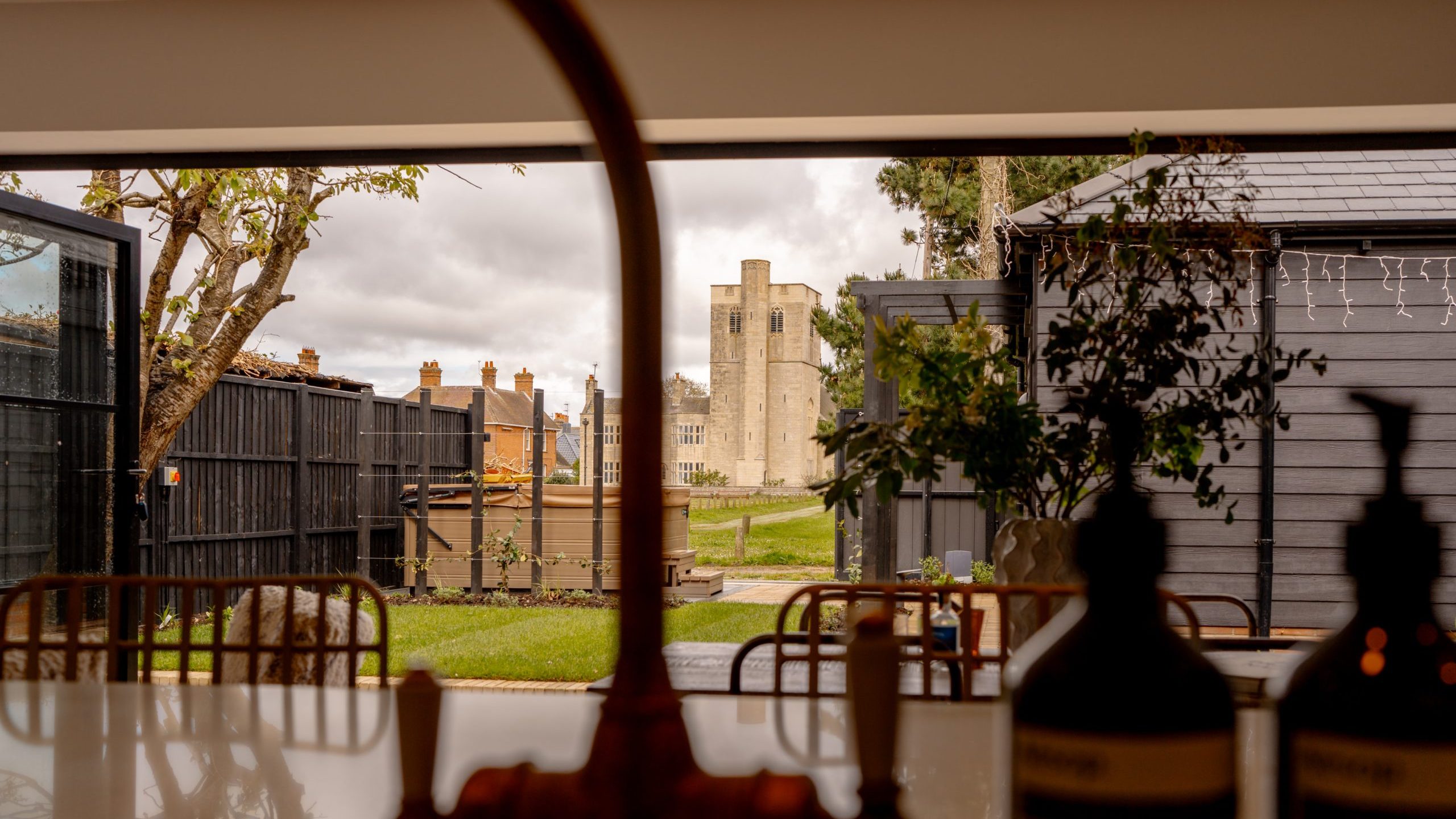 View from inside a cafe, looking out through a window framed by bottles at the bottom, showcasing a distant historic stone castle tower, surrounded by greenery and a cloudy sky.