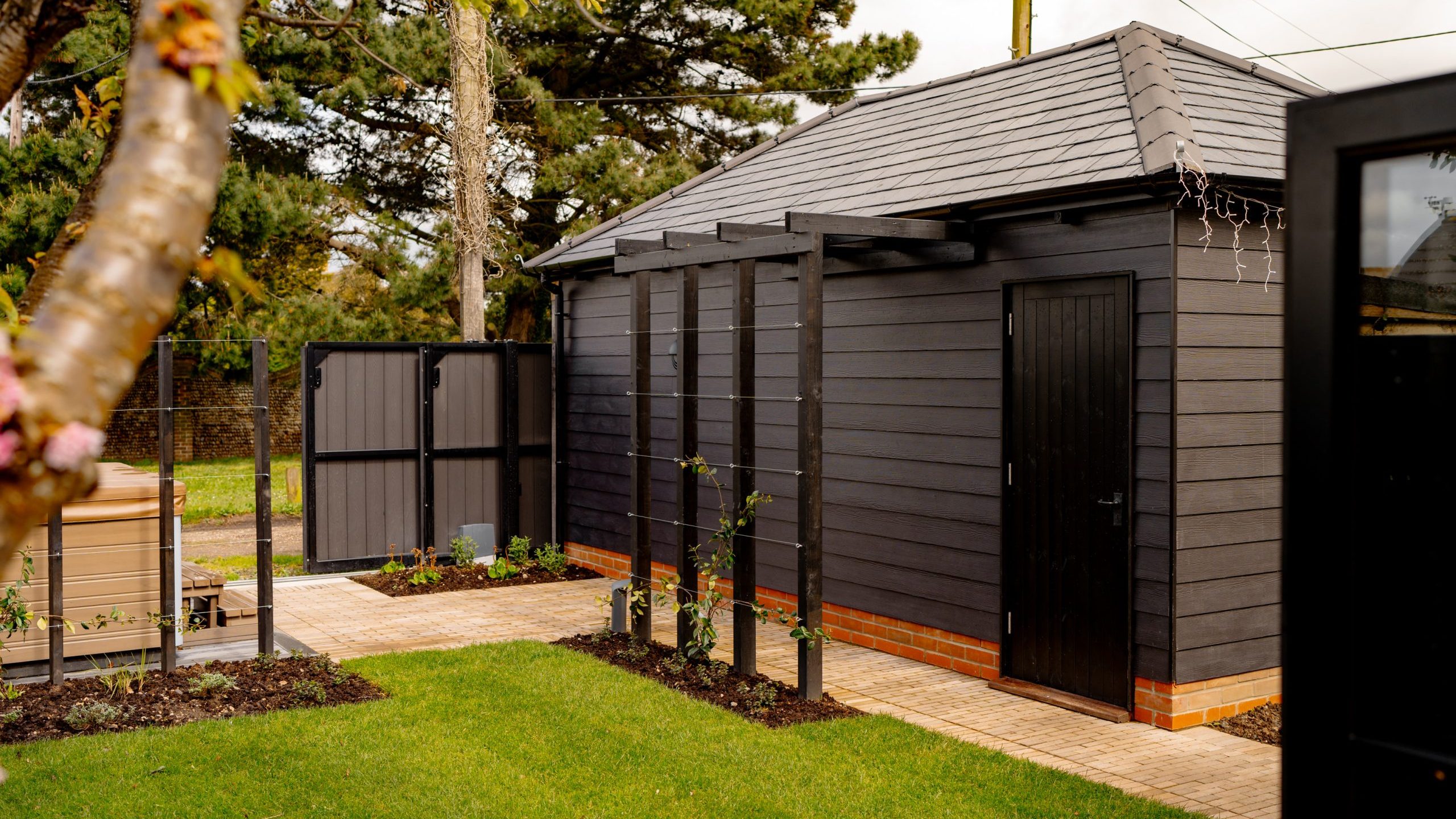 A modern black garden shed with a sloping roof, surrounded by a neatly manicured lawn and a brick pathway. a decorative plant and a small fence are visible, with the focus slightly blurred in the foreground.