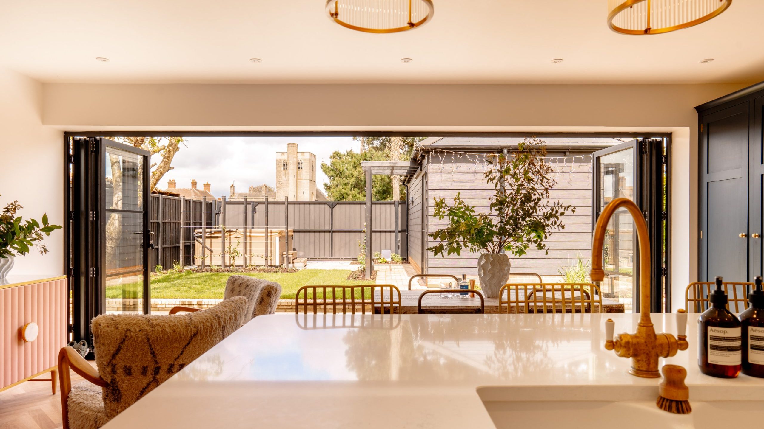 A modern kitchen with an open layout featuring large, accordion-style doors leading to a sunny backyard. the room has sleek countertops, warm lighting fixtures, and a cozy sitting area.