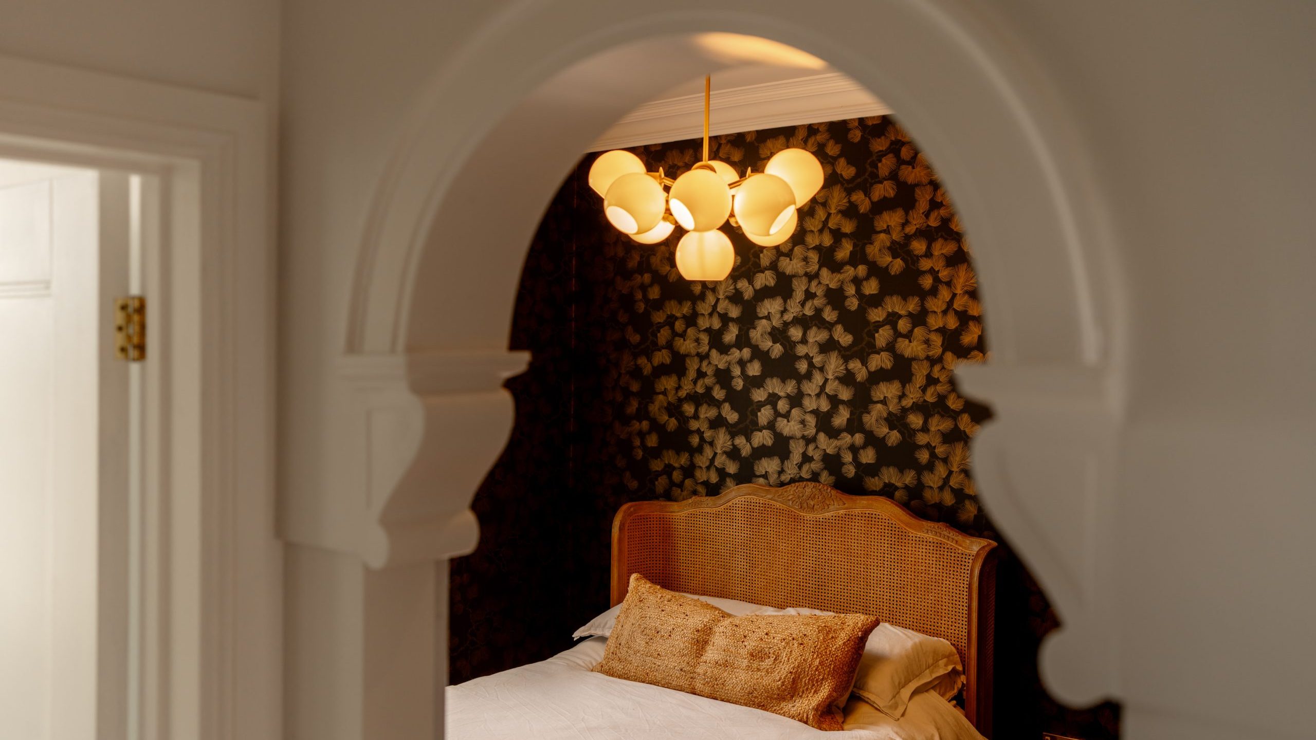 View through an arched doorway of a cozy bedroom featuring a rattan headboard, decorative floral wallpaper, and a modern hanging light fixture. a white door is partially visible on the left.