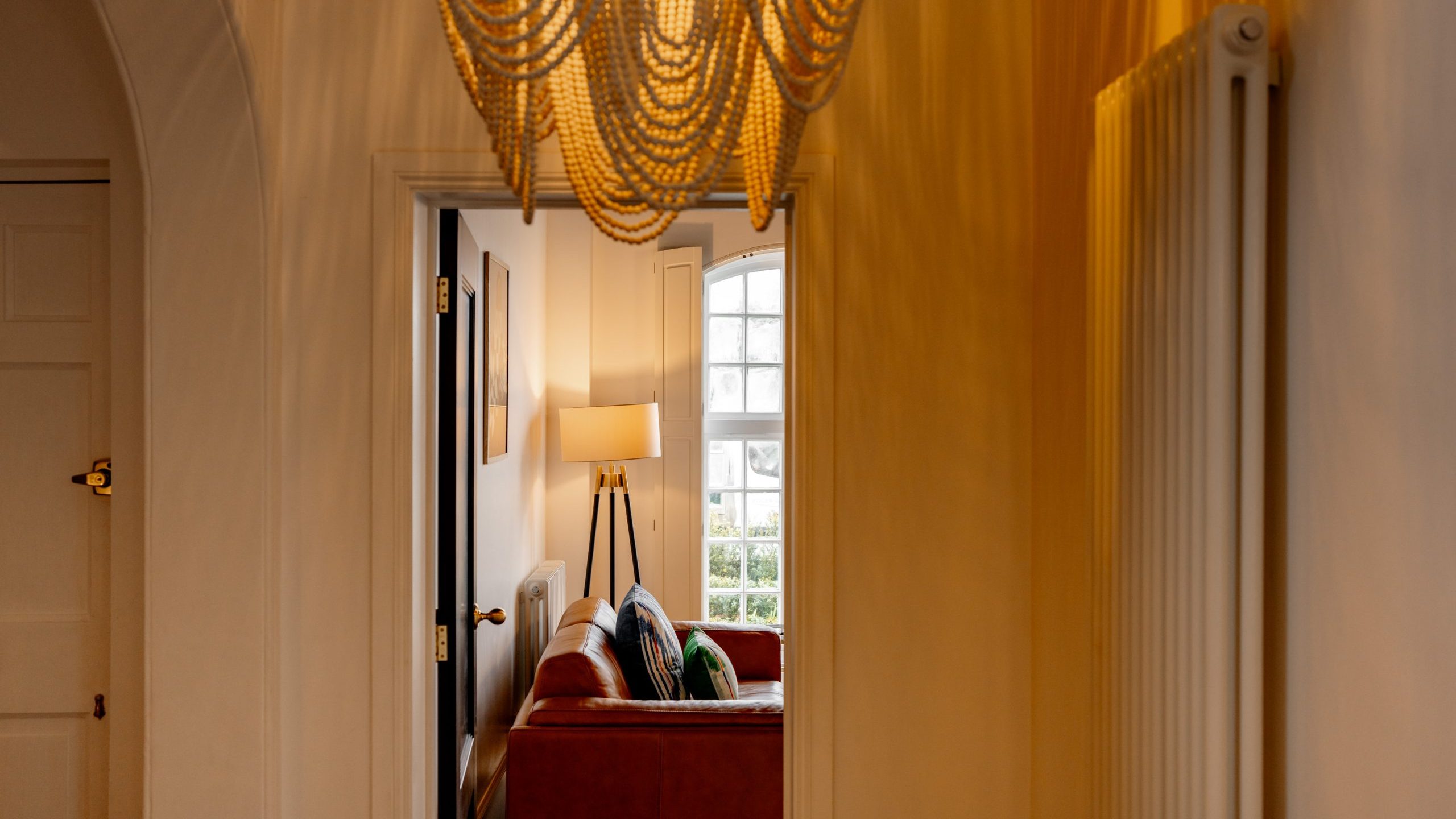Interior view of a cozy living room seen through an archway, featuring an elegant chandelier, warm yellow curtains, and a sofa with a green throw pillow under a lamp by a window.