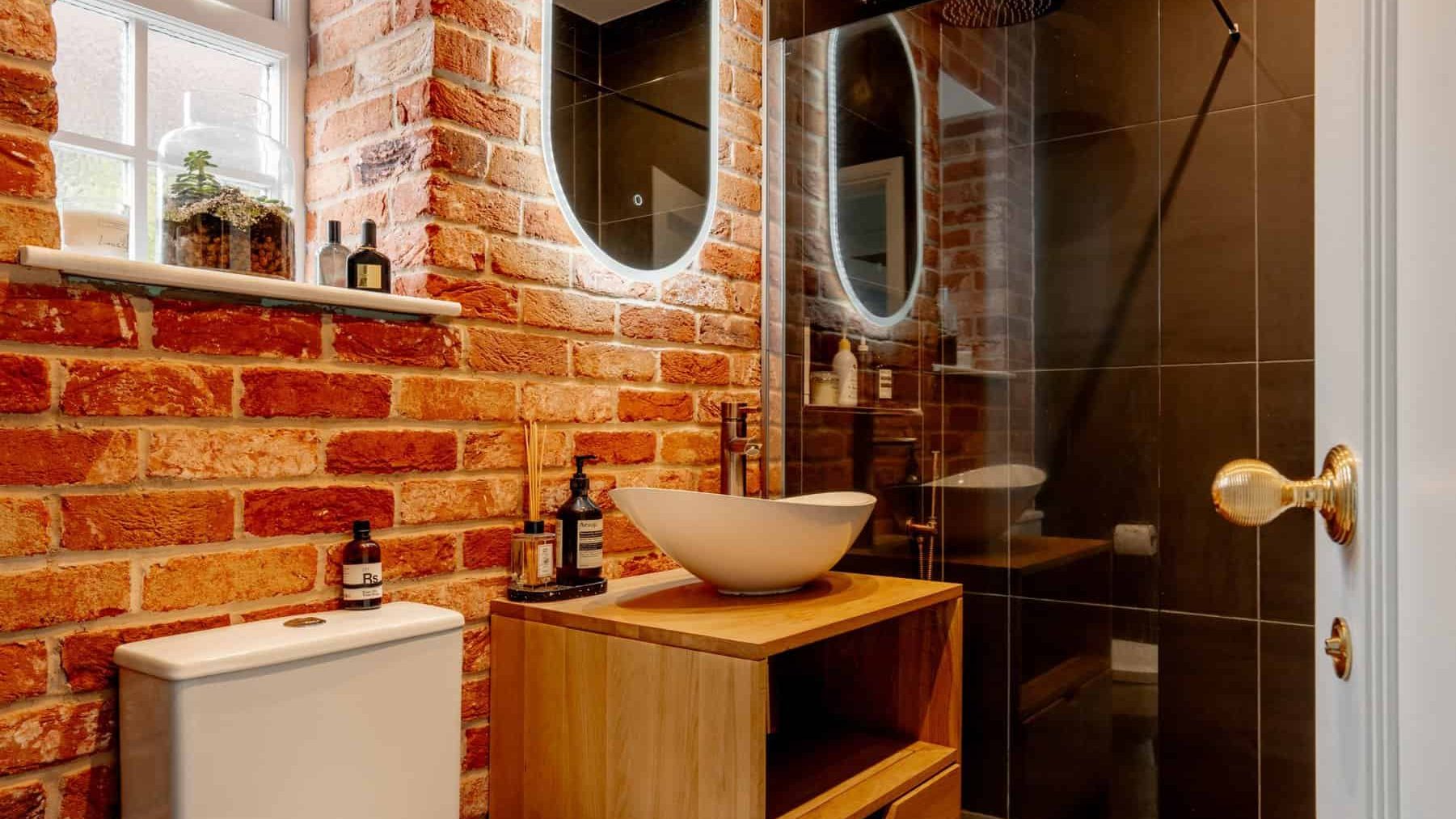 A modern bathroom with exposed brick walls, featuring a glass shower, wooden vanity with a vessel sink, and a round mirror with backlighting. natural light streams in through a small window.