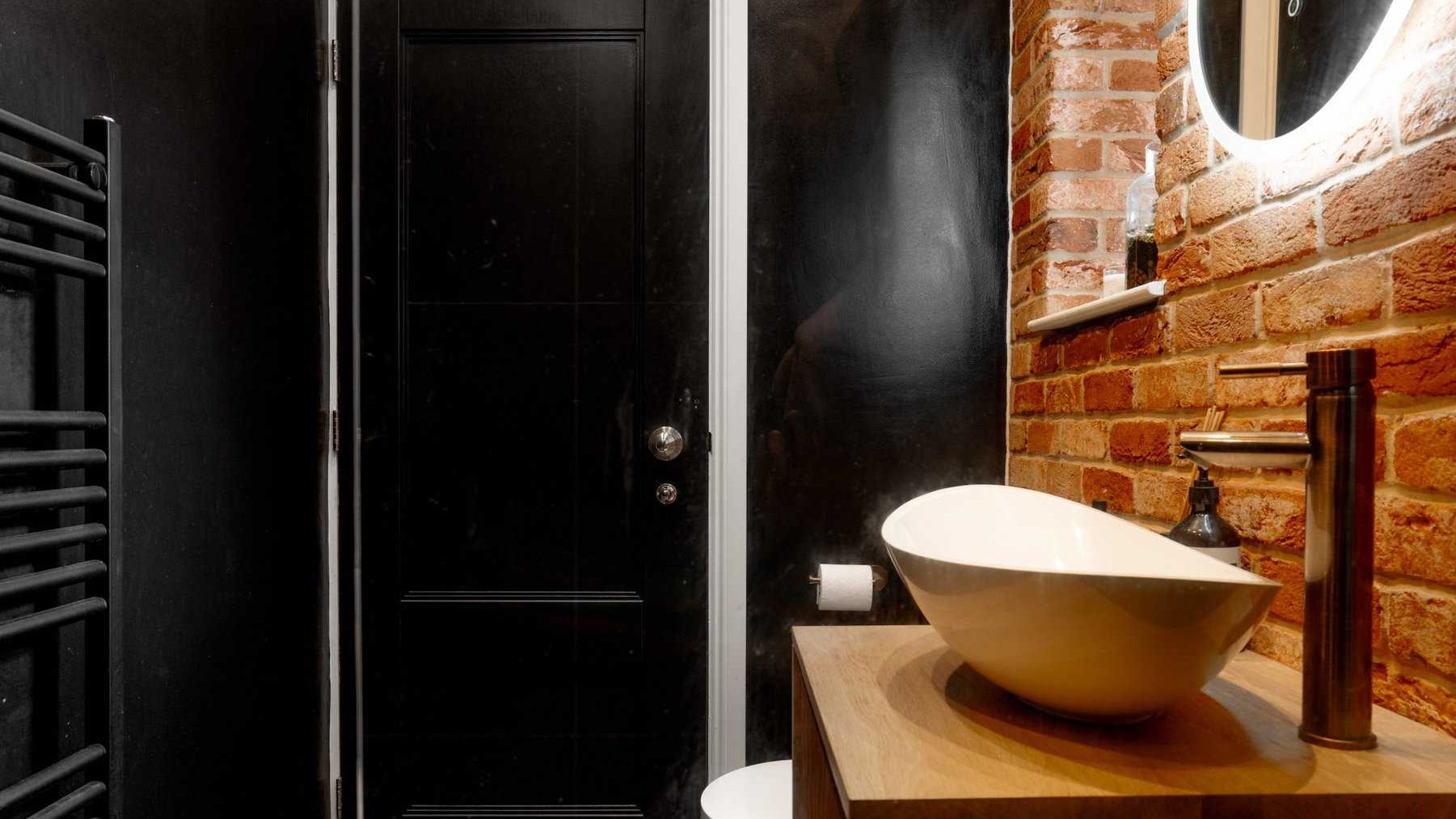 Modern bathroom featuring a black door, exposed brick wall, and a wooden countertop with a white oval basin. there's a round mirror above the basin and a heated towel rail on the left.