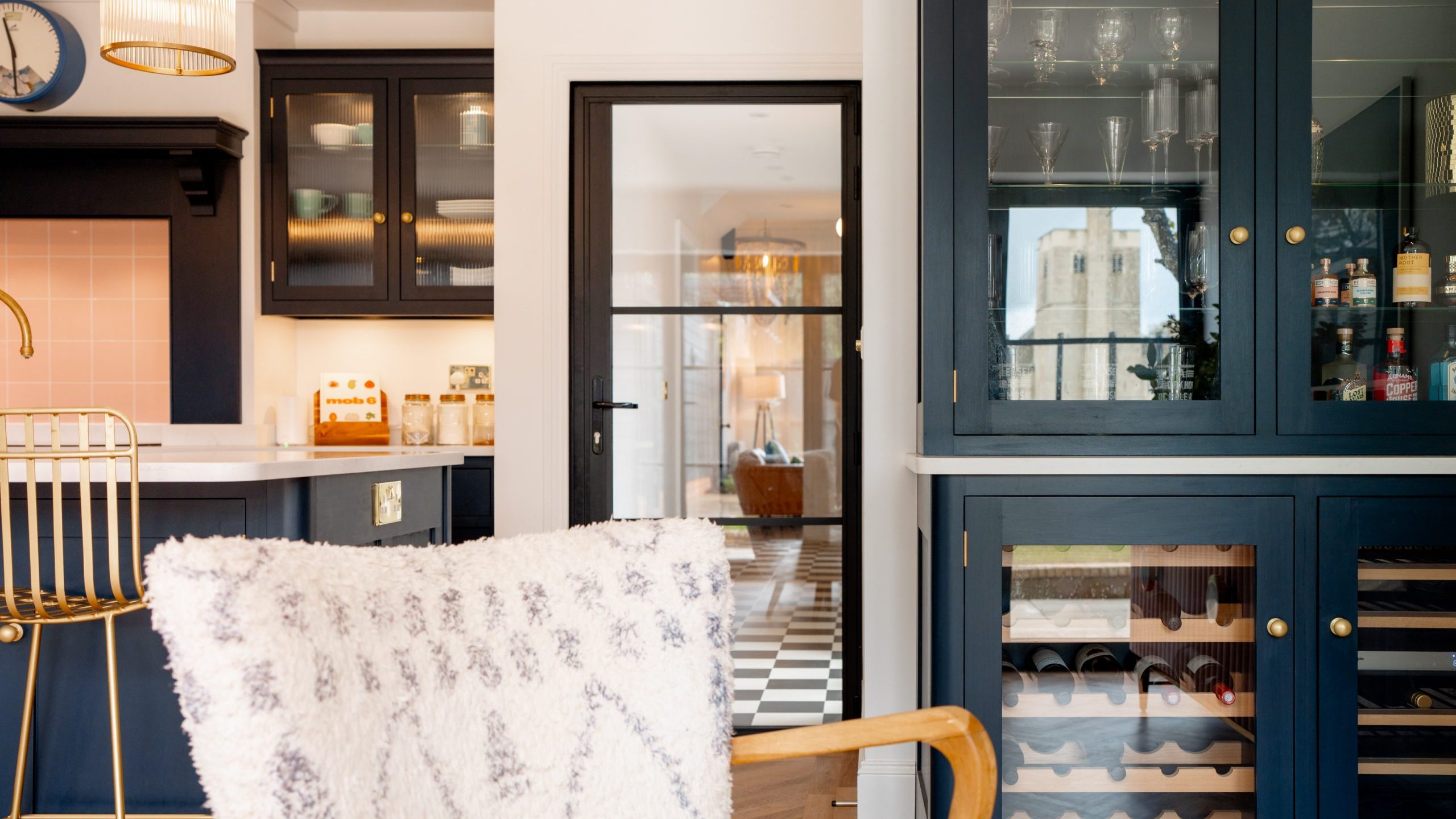 A stylish kitchen with dark blue cabinetry and a gold bar chair in the foreground. glass cabinet doors display glassware, adjacent to a built-in wine fridge. a doorway leads to another room with a checkered floor.