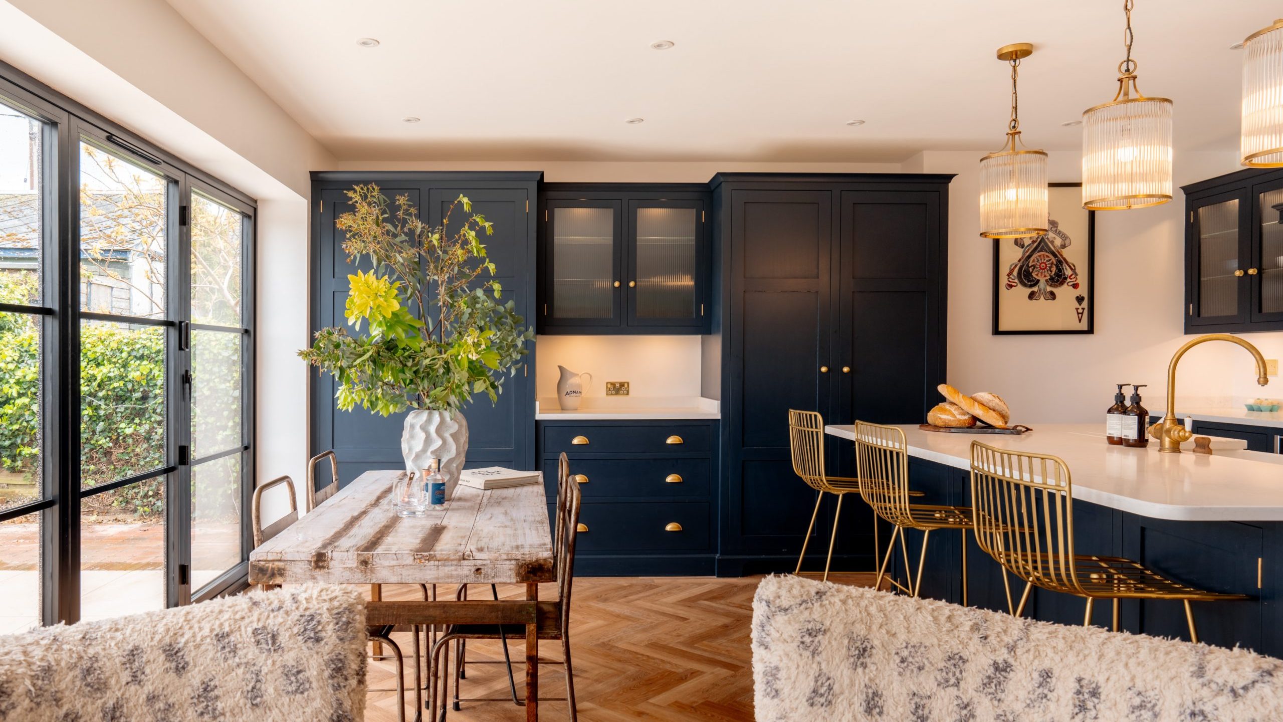 A modern kitchen with deep blue cabinetry, gold handles, and integrated appliances. a wooden dining table with textured chairs is adjacent to the kitchen island. sunlight streams through large windows.