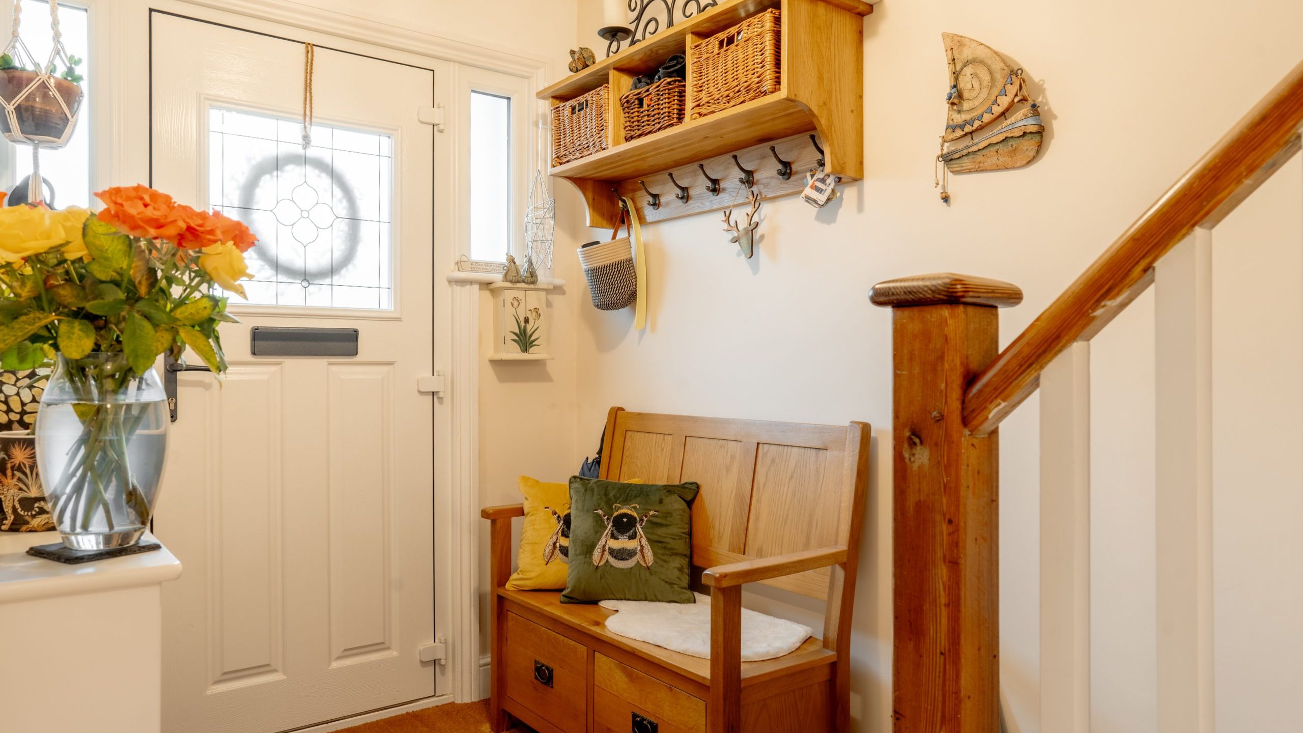 A cozy entryway featuring a wooden bench with storage underneath, a cushion with an owl design, and a bouquet of orange roses. the wall is decorated with rustic shelves, a hat, and wicker baskets.