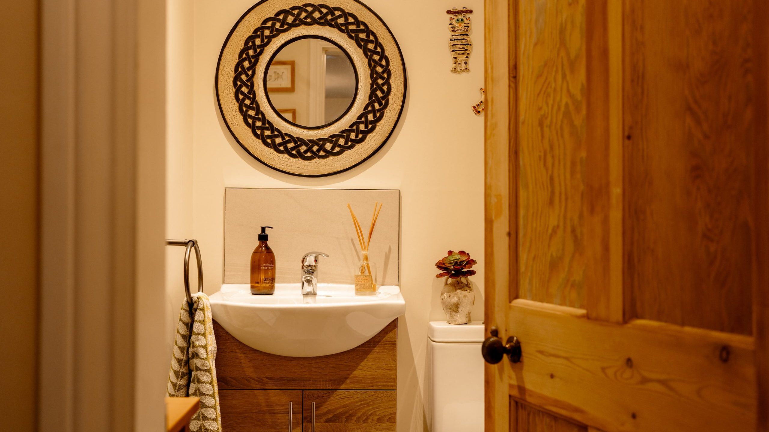 A cozy bathroom with a wooden door partly open, revealing a wooden vanity topped with a white sink, stylish soap dispenser, and decorative items beside a circular mirror with a black frame.