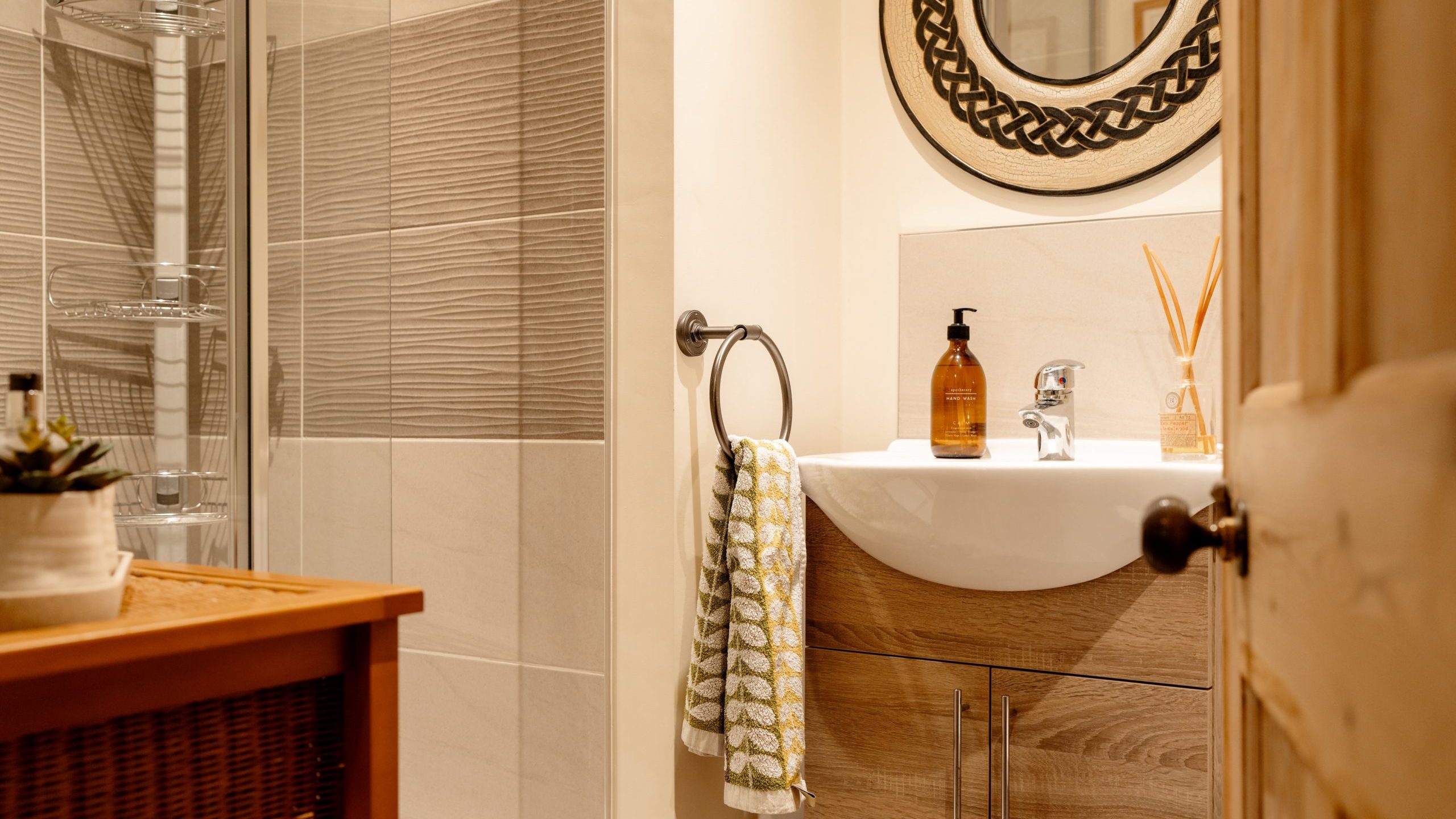 A cozy, well-lit bathroom featuring a round mirror, a stylish sink with wooden vanity, beige tiles, and neatly arranged toiletries and towels.