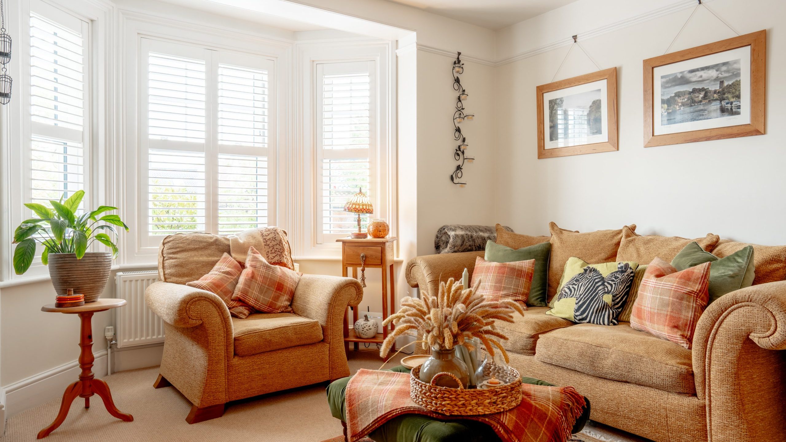 A cozy living room with natural light, featuring two plush armchairs, a sofa filled with cushions, a coffee table with a plant centerpiece, and framed pictures above.