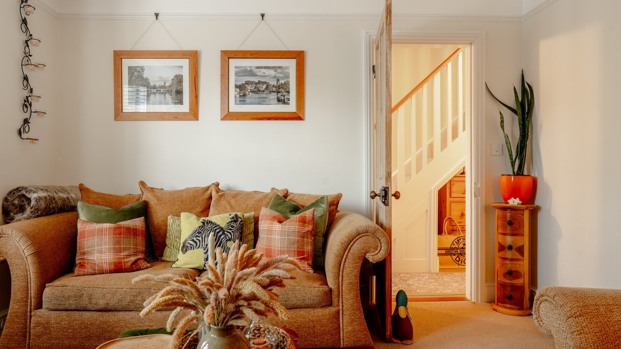 Cozy living room with a plush sofa adorned with colorful pillows, framed pictures on the wall, a vibrant rug, and a small side cabinet with a plant next to an open door.
