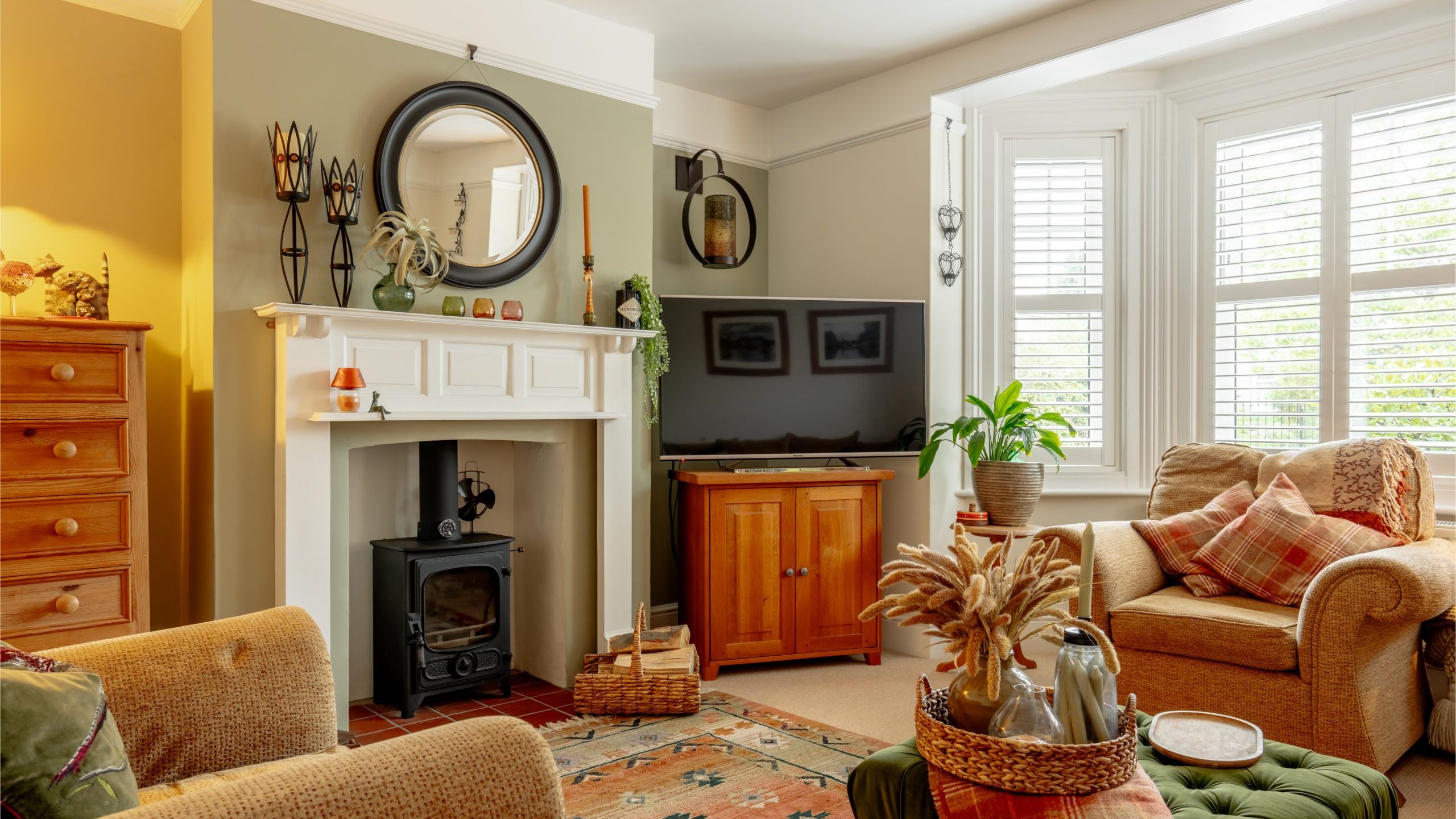 Cozy living room with a white fireplace, wooden furniture, and a plush sofa near large windows with blinds. decor includes a tv, plants, and a patterned rug.