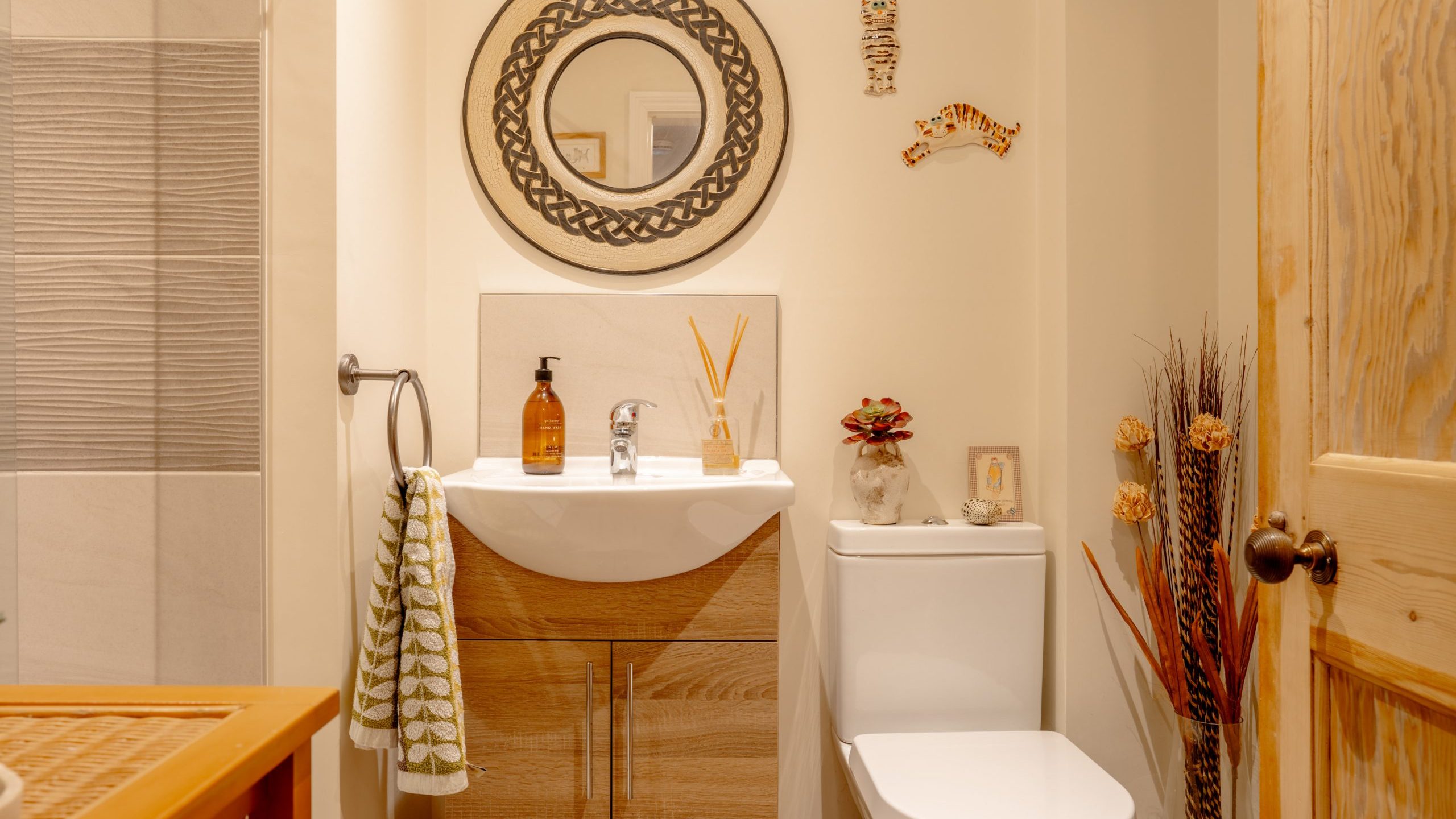 A cozy bathroom featuring a white sink, mirror with circular frame, toilet, and decorative wooden elements, complemented by warm lighting and beige tiles.