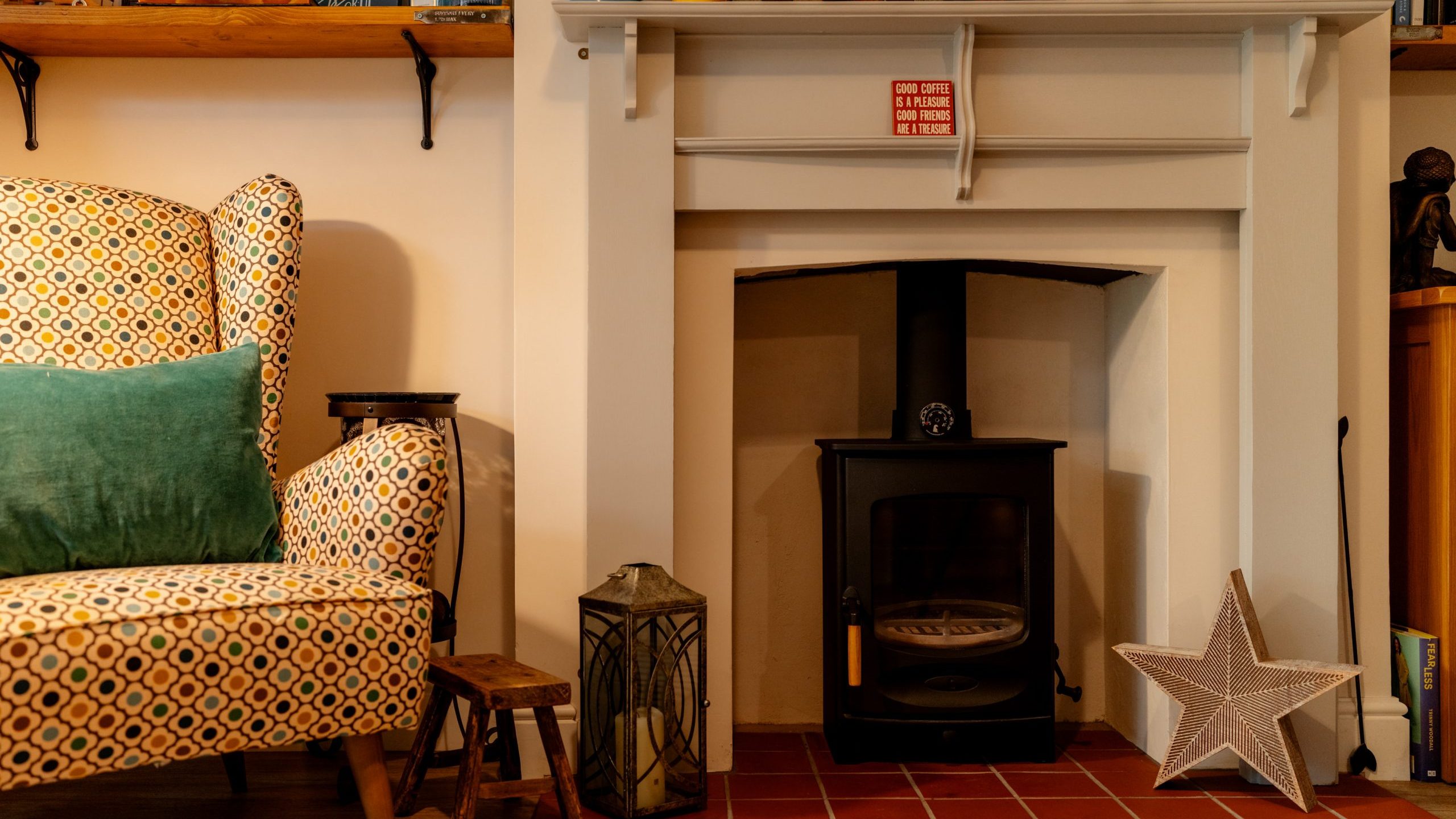 Cozy living room corner featuring a colorful patterned armchair next to a brick-lined fireplace with a wood-burning stove, decorative star, and lantern on a tiled hearth.