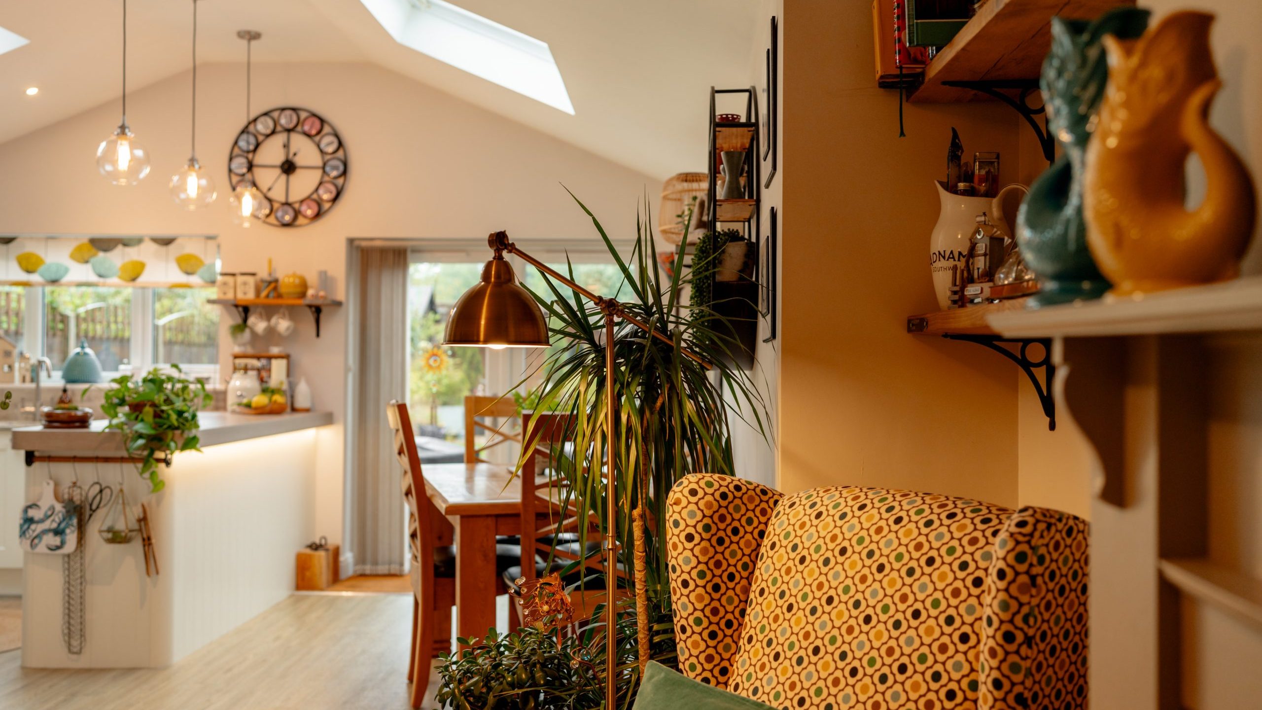 Cozy and warmly lit living room with a vibrant yellow patterned armchair, various houseplants, and decorative items. a well-equipped kitchen is visible in the background under a vaulted ceiling.