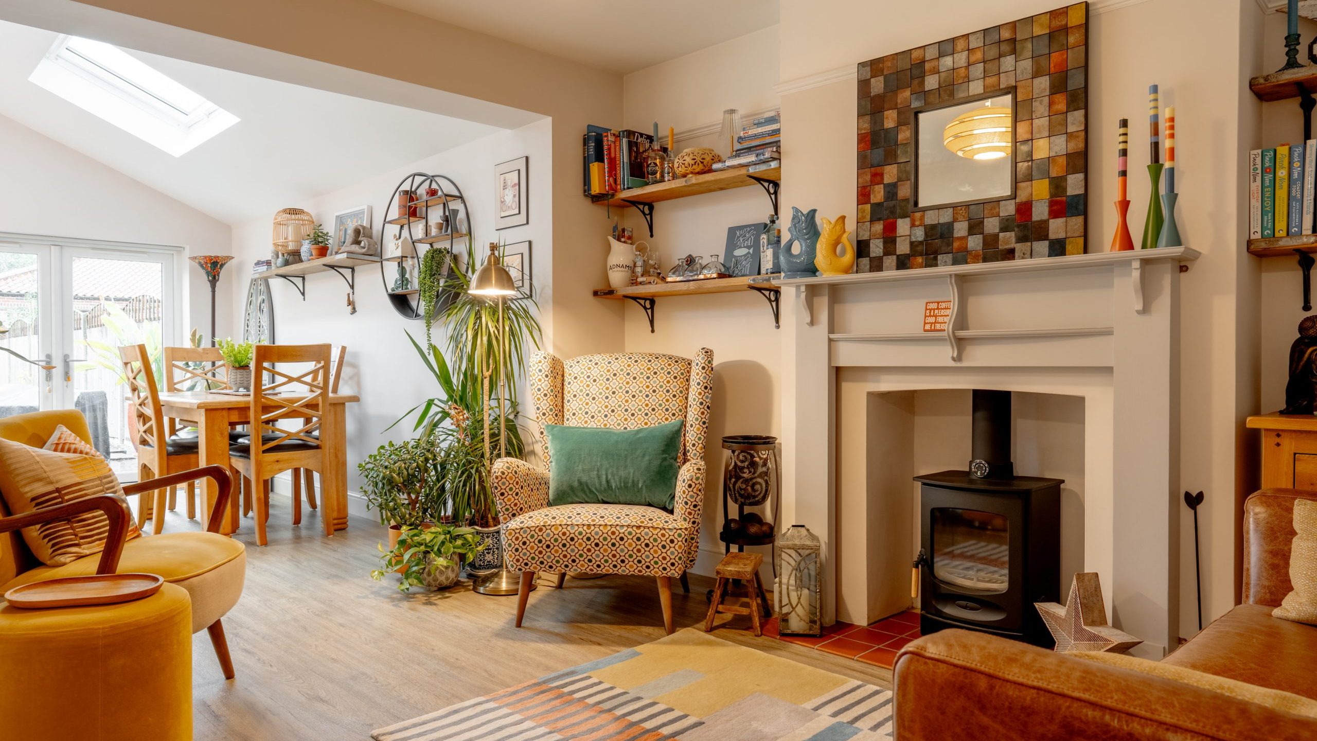A cozy living room featuring a skylight, patterned armchair, a fireplace, and various houseplants. there are shelves with books and décor, and a colorful rug on the floor.