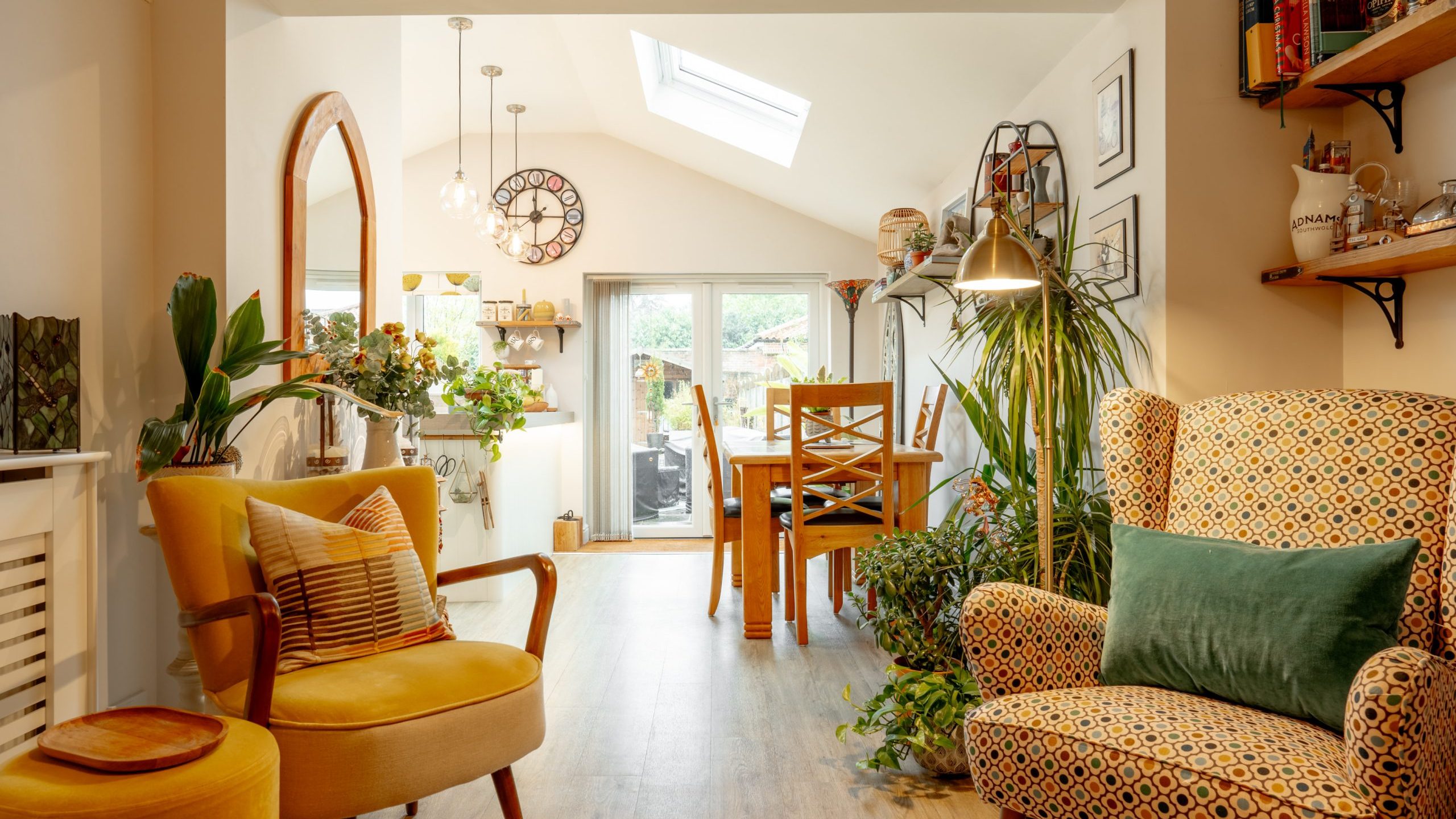 Cozy, well-lit living room with a mix of modern and vintage decor featuring vibrant patterned chairs, a variety of potted plants, and a wall of books. skylights and large glass doors brighten the space.