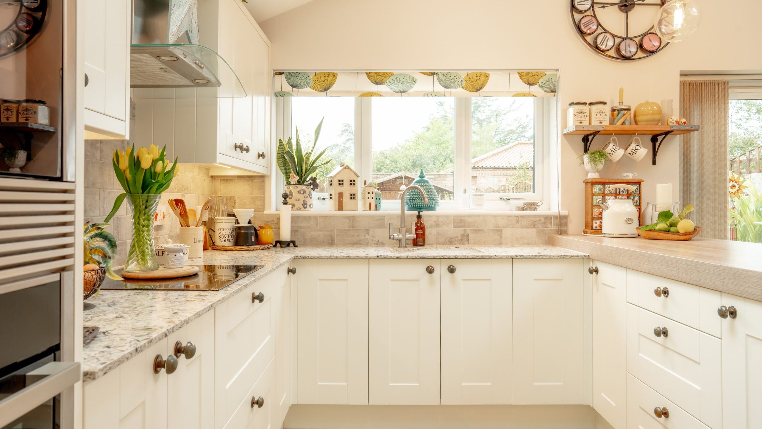 Bright, cozy kitchen interior featuring white cabinets, granite countertops, and a large window with a view of greenery outside. decor includes a wall clock, tulips, and neatly arranged kitchenware.