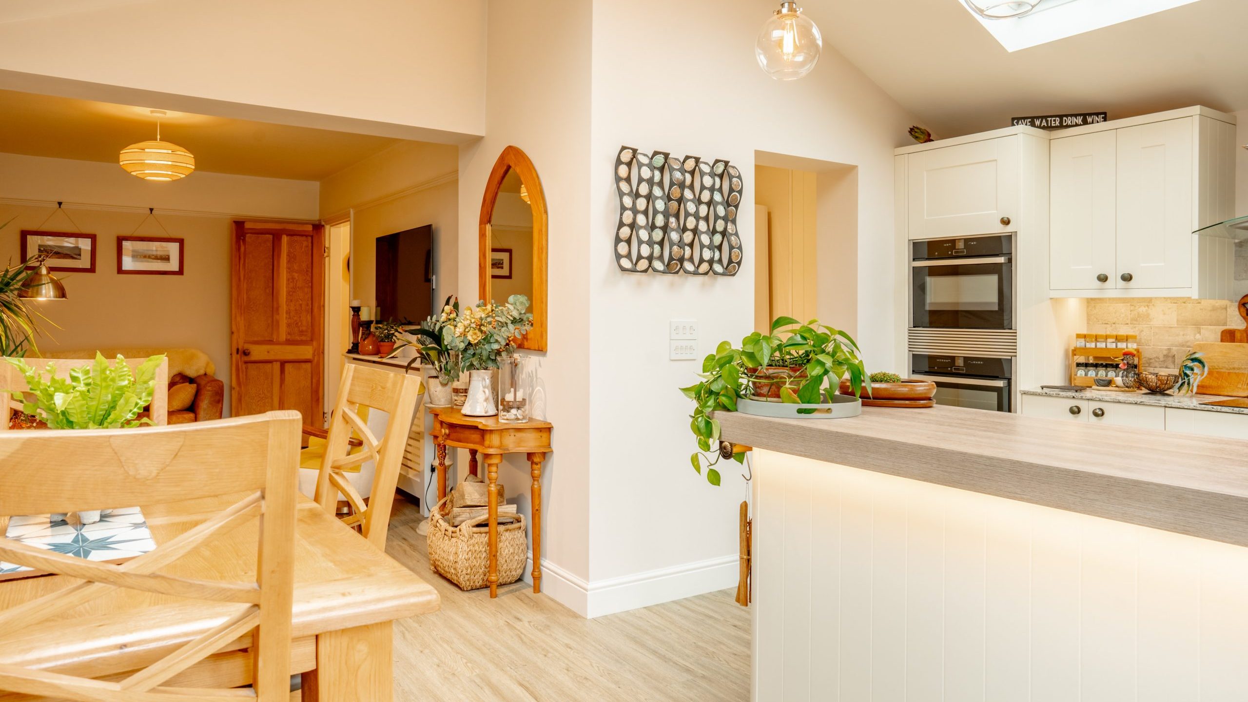 A cozy, well-lit kitchen featuring white cabinetry, stainless steel appliances, a central island, and a dining area with wooden furniture. decor includes plants and hanging light fixtures.