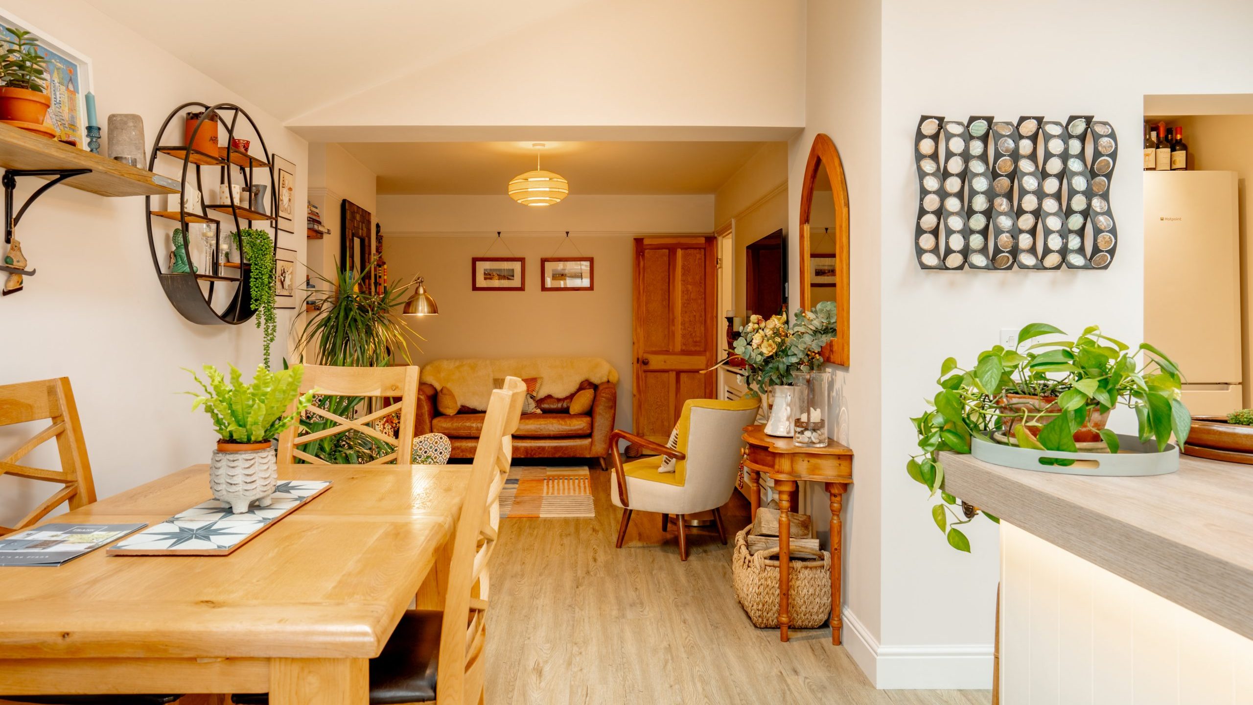 A cozy, well-lit dining room with a wooden table and chairs, decorative plants, and a view into a living room with a yellow sofa and artwork.