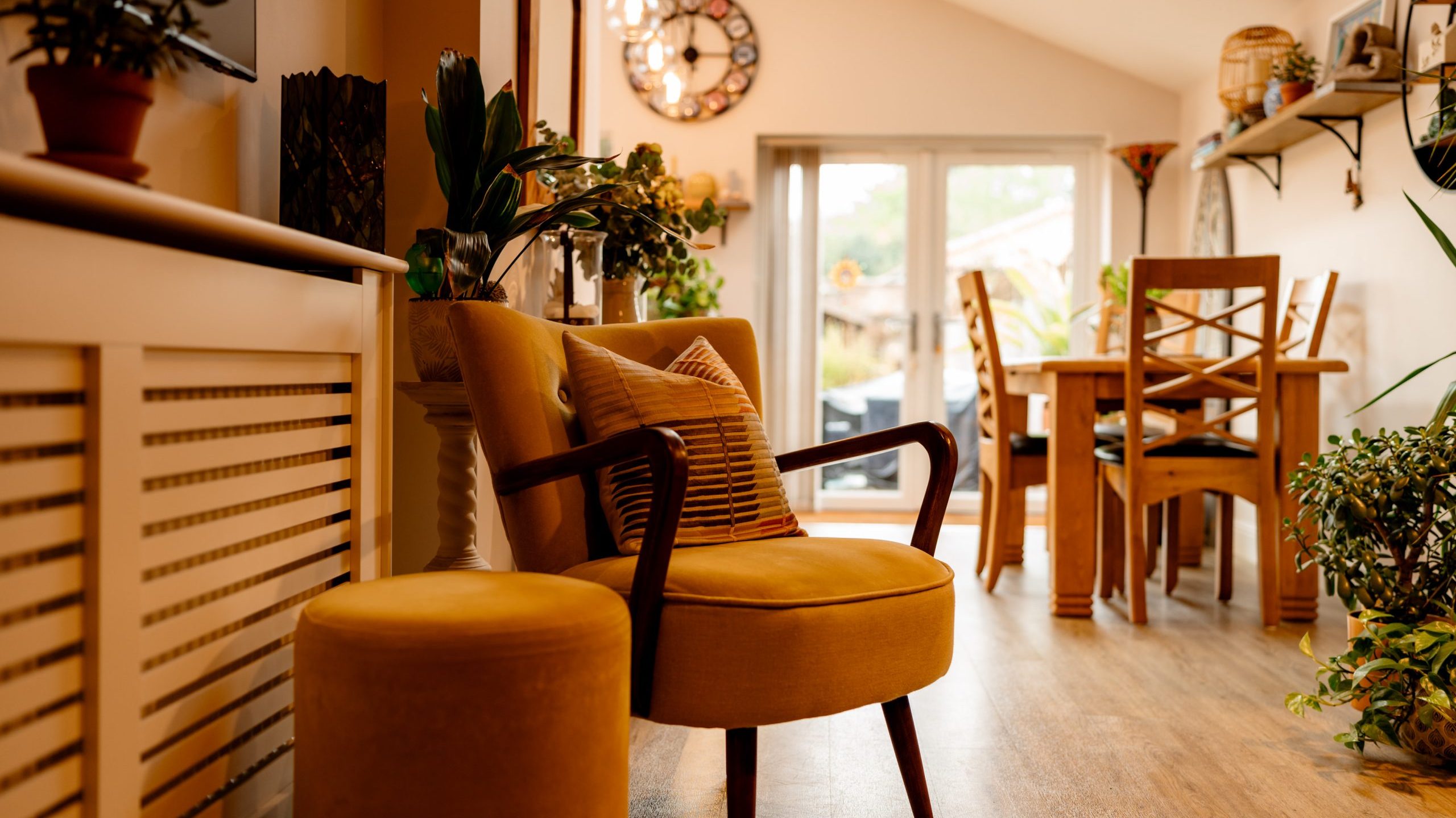 A cozy living room with plants and wooden furniture, featuring a stylish yellow armchair with a striped cushion, and sunlight streaming through open doors.
