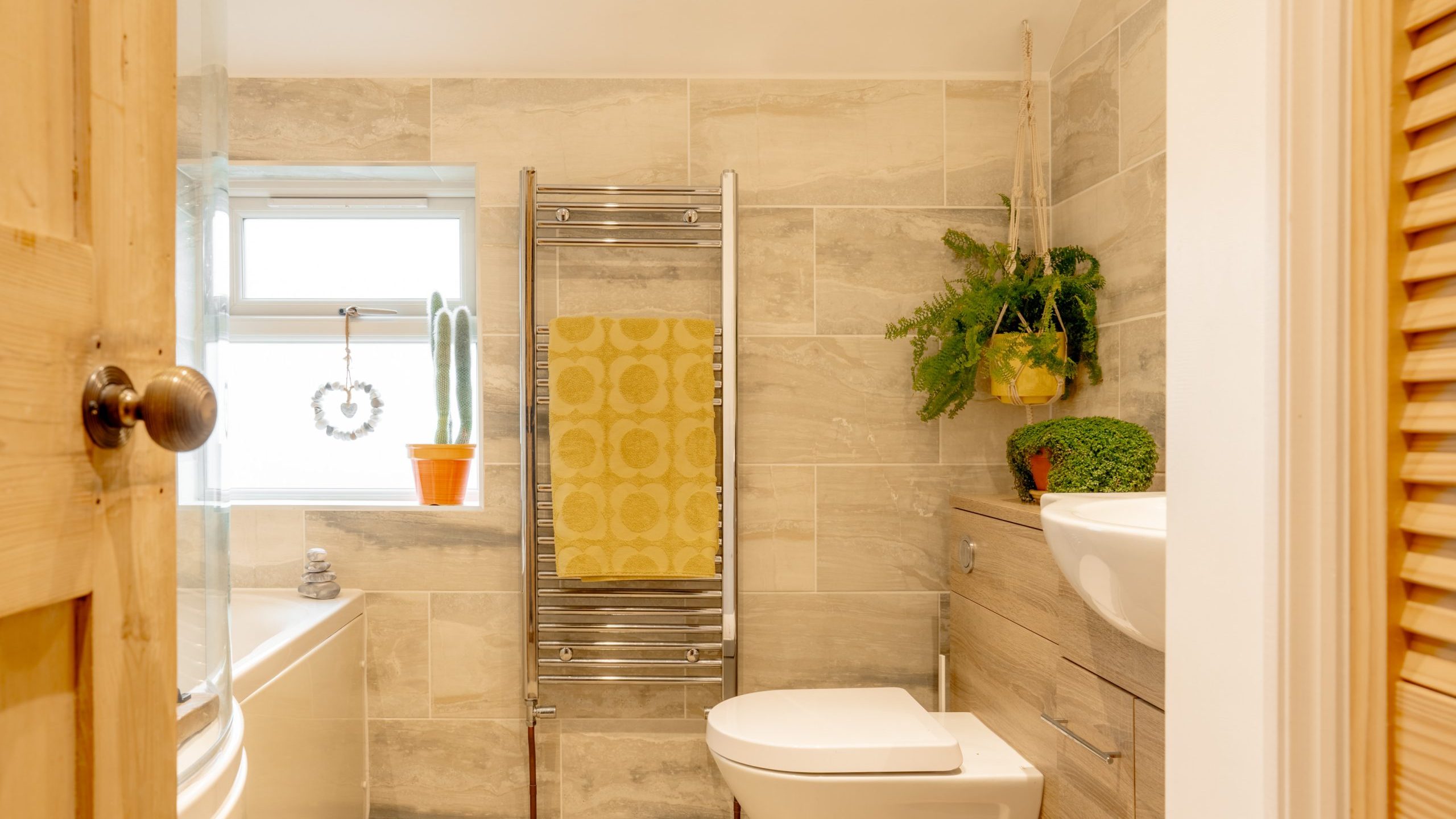 Modern bathroom interior with beige tile walls, heated towel rack holding a yellow towel, a toilet, and a wooden sink cabinet. a window allows natural light to enter.