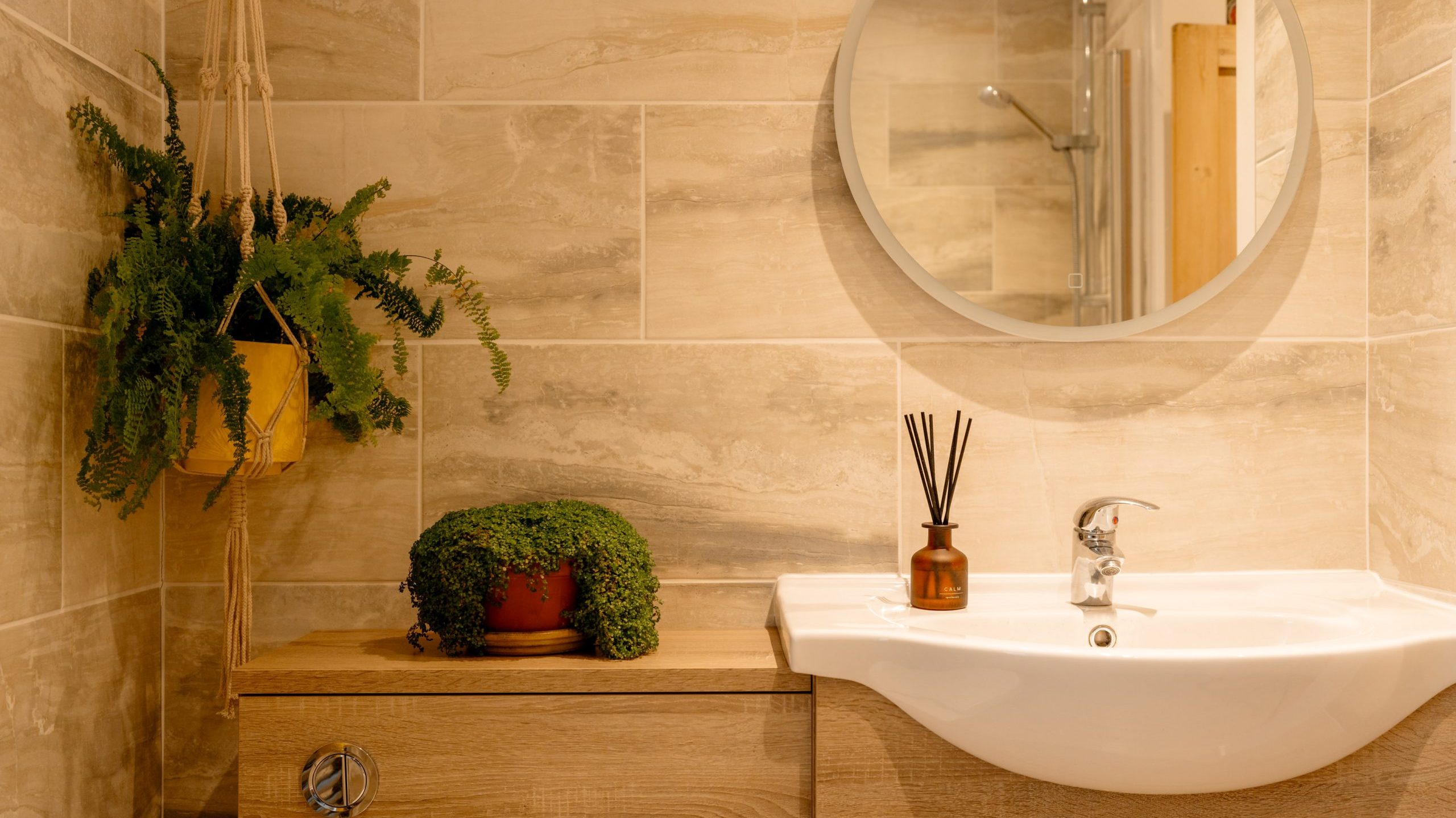 A modern bathroom with warm lighting, featuring a wooden vanity, white basin, round mirror, and plants as decoration, with a visible shower area in the reflection.