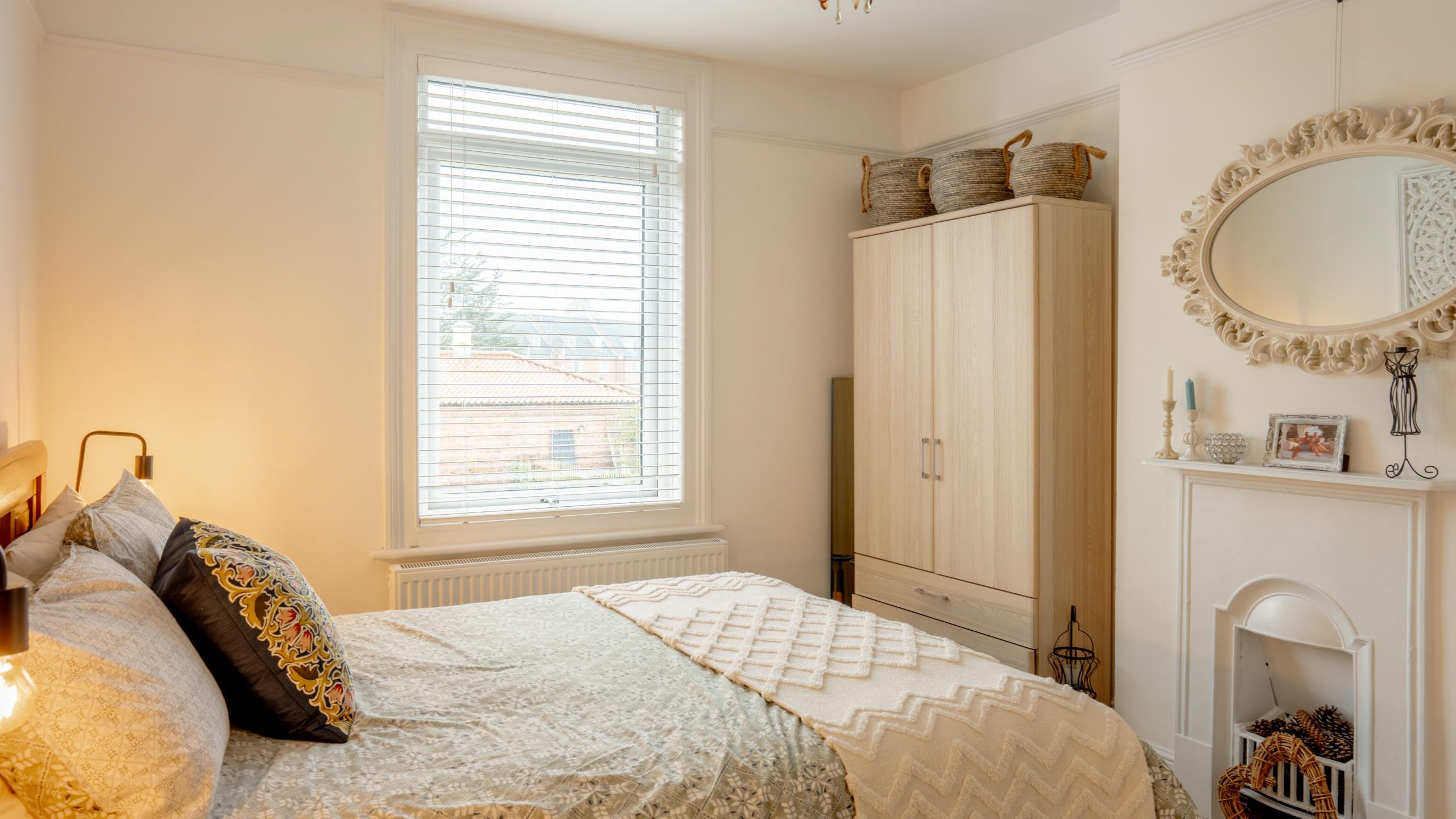 Cozy bedroom featuring a double bed with patterned bedding, a white wardrobe, and a mirror above a decorative fireplace, with a window offering natural light.