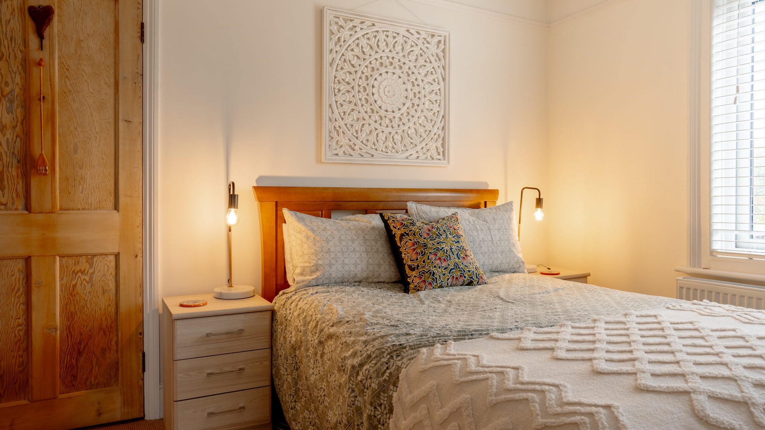 A cozy bedroom with a wooden double bed covered in a quilted blanket, flanked by lit bedside lamps, a decorative white wall art above, and a wooden door to the left.
