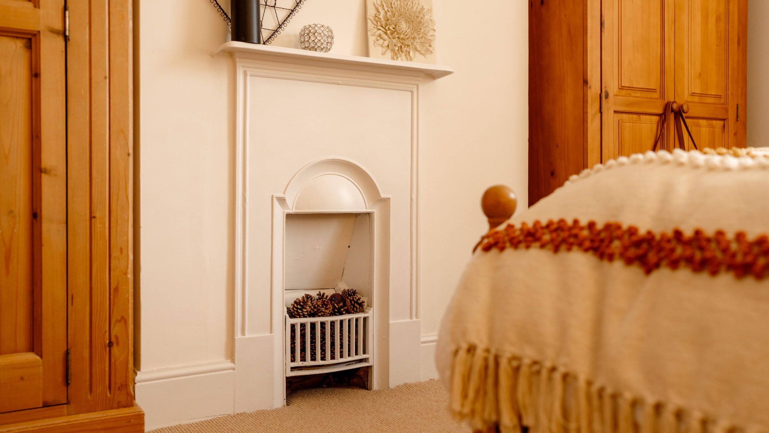 A cozy room corner featuring a white decorative fireplace with a collection of pine cones, flanked by a wooden door and a wall art, with a part of a bed draped with a fringed blanket visible in the foreground.